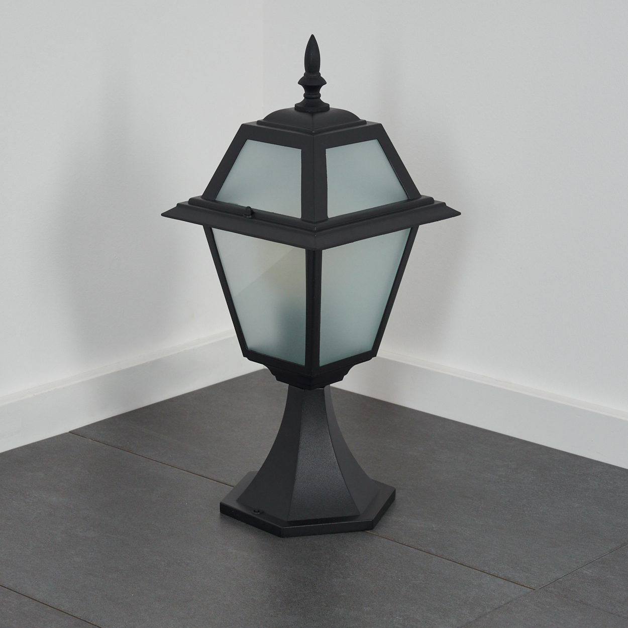 Eine schwarze Außenlampe steht in einer Ecke. Sie hat einen sechseckigen Sockel, einen Schirm aus Milchglas und eine spitze Spitze auf grauen Fliesen.