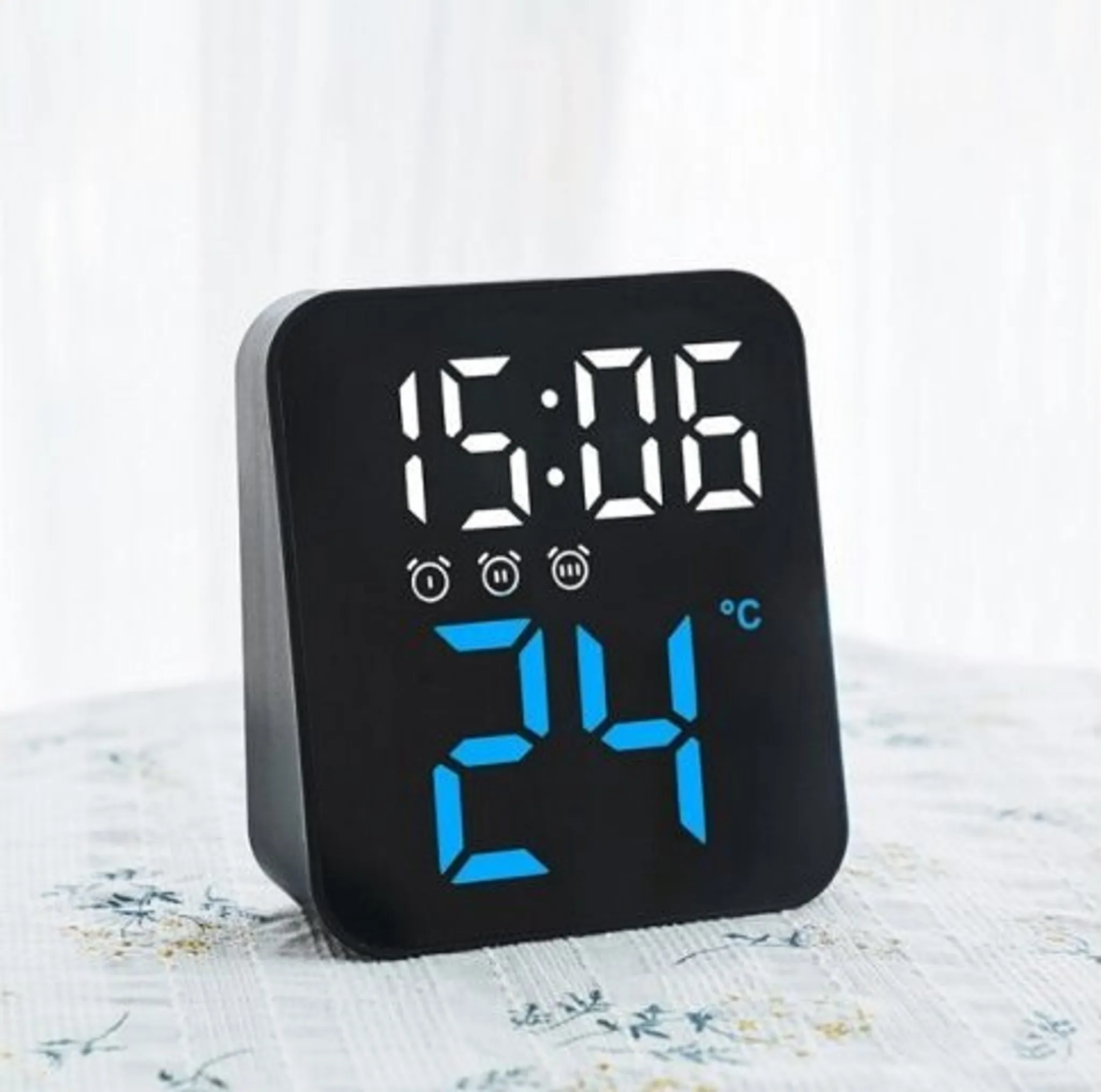 Eine schwarze Digitaluhr zeigt 15:06 und 24°C auf einer gemusterten Oberfläche. Weißer Hintergrund.