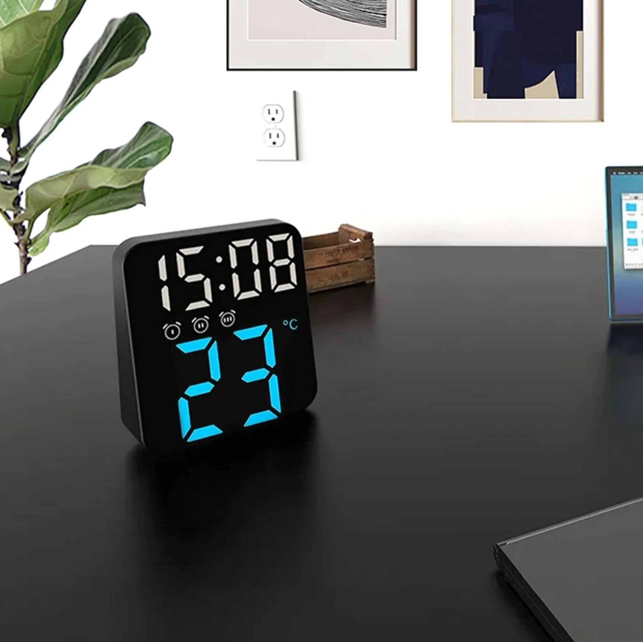 Eine Digitaluhr zeigt 15:08 und 23 Grad an. Ein Laptop, Wandkunst und eine Pflanze sind ebenfalls auf dunkler Oberfläche sichtbar.
