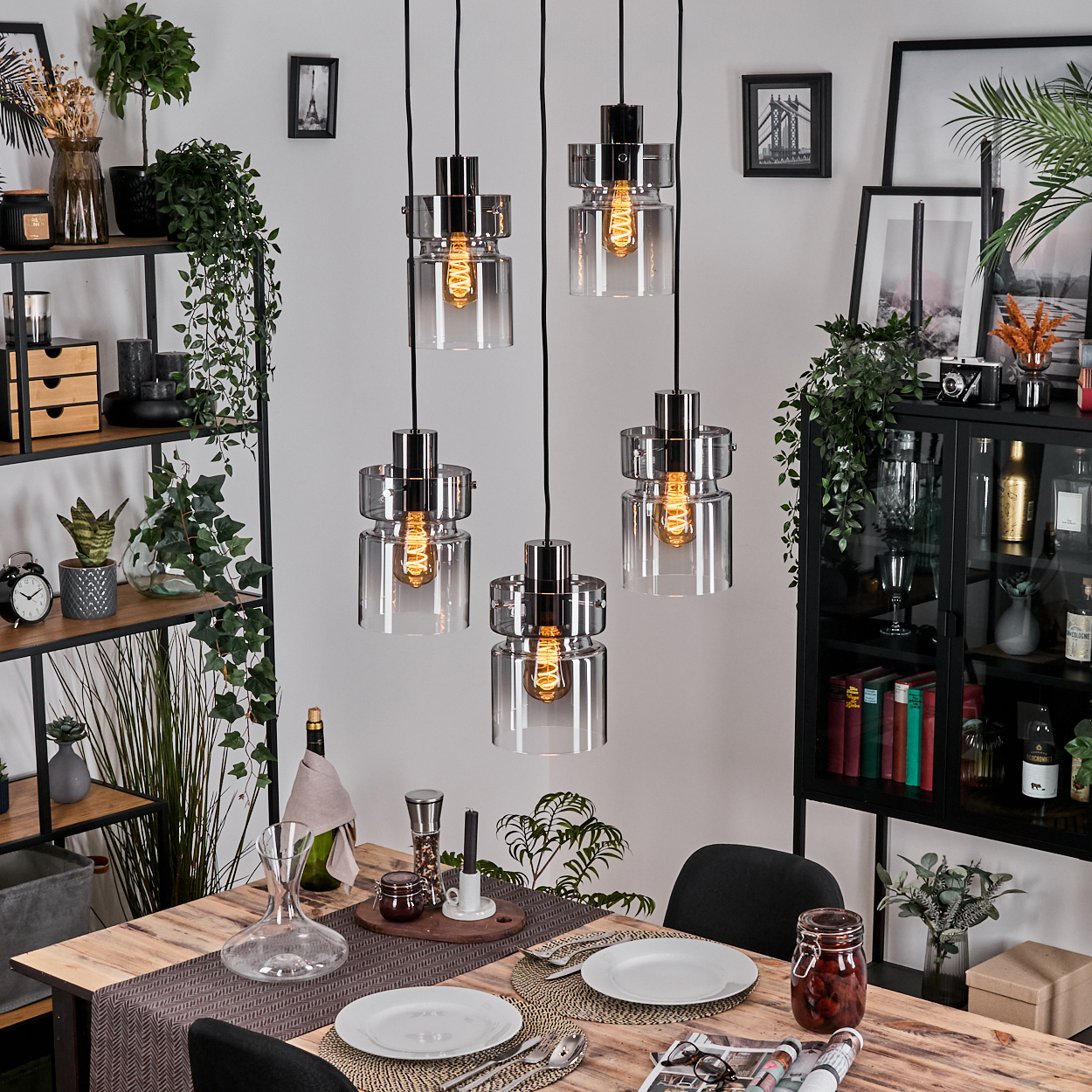 Esszimmer mit Holztisch, Hängelampen, Pflanzen und Dekorationsartikeln. Tisch für eine Mahlzeit gedeckt. Schwarze Stühle.