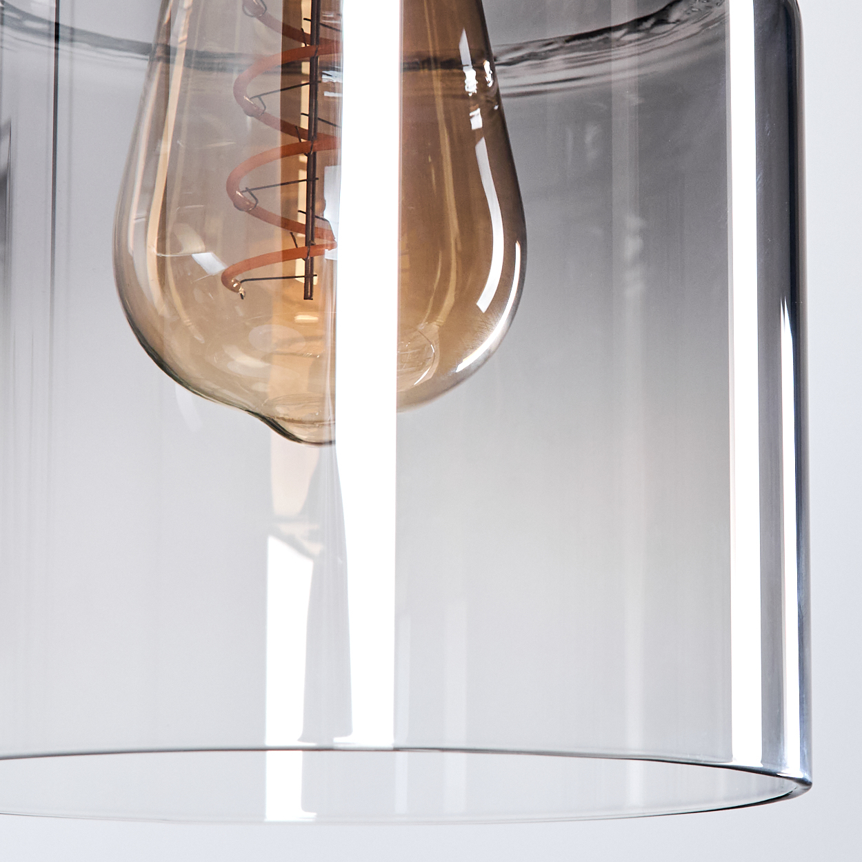 Nahaufnahme einer Glühbirne in einem Glaszylinder. Die Birne hat einen Spiralfaden. Das Glas hat einen Farbverlauf von klar zu grau.