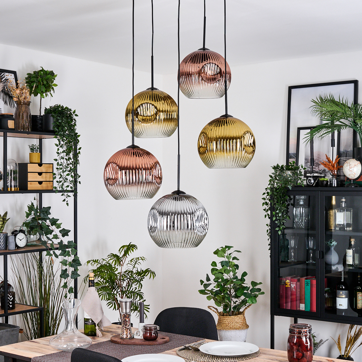 Esszimmer mit Hängelampen, Pflanzen und Esstisch. Lampen haben goldene, kupferne und silberne Schirme. Der Tisch ist gedeckt.