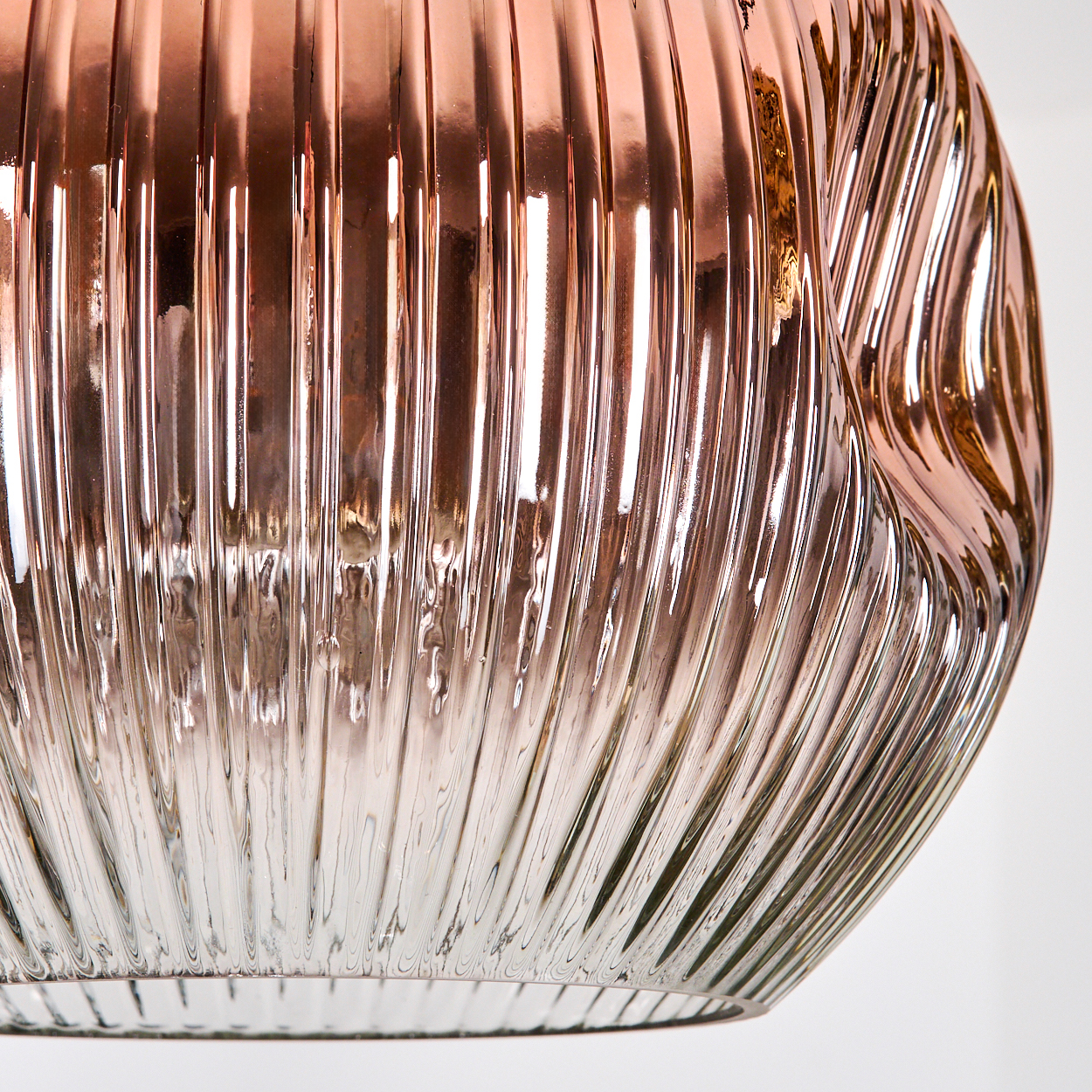Nahaufnahme eines Glasobjekts mit vertikalen Rippen. Das Glas hat einen Farbverlauf von Roségold zu klar, mit Lichtreflexionen auf weißem Hintergrund.