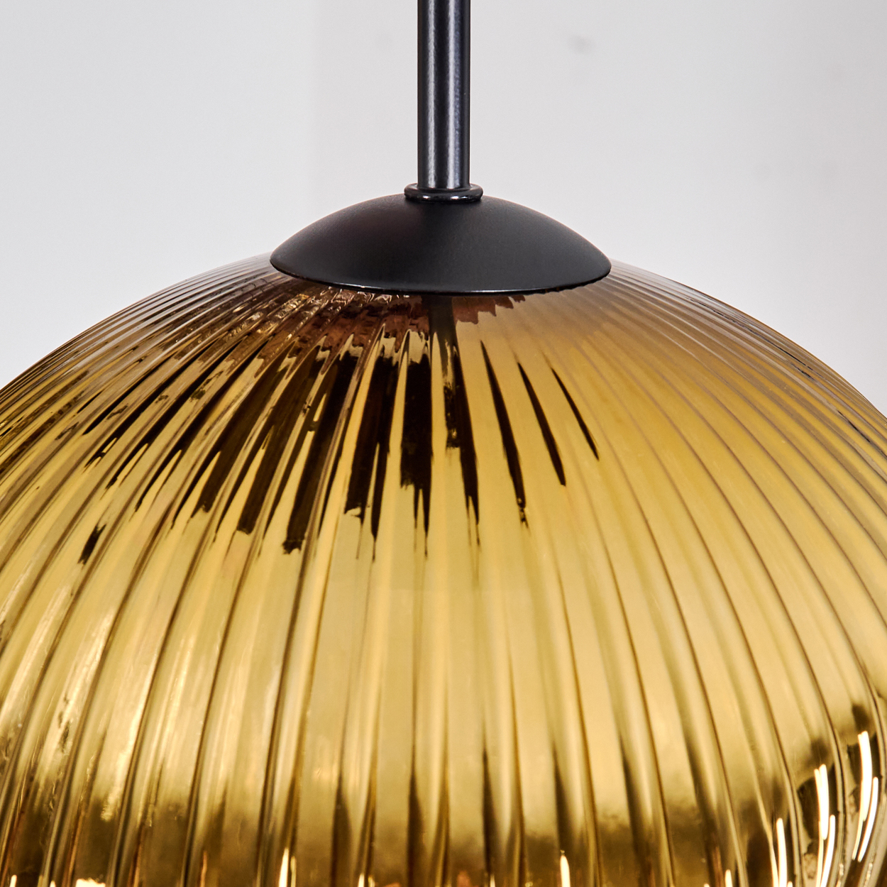 Nahaufnahme eines goldfarbenen, gerippten Glasschirms, der an einem schwarzen Stiel hängt. Der Hintergrund ist schlicht weiß.
