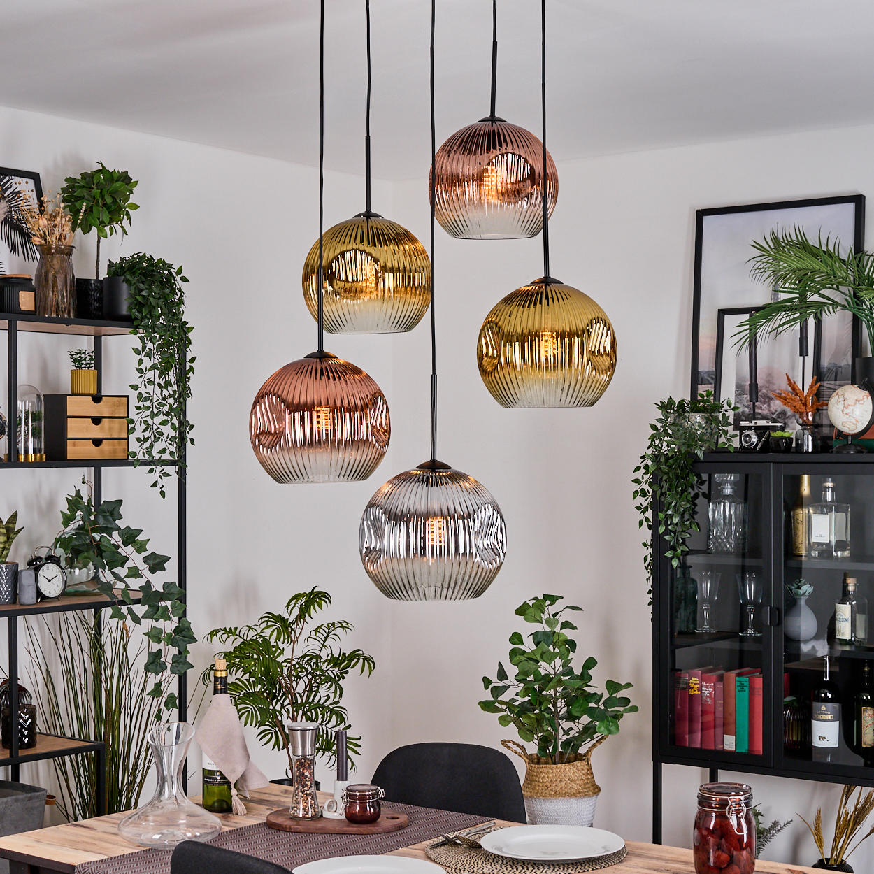 Esszimmer mit hängenden Kugelleuchten in verschiedenen Metallic-Tönen. Tisch mit Tellern und Dekoration. Pflanzen und Regale im Hintergrund.