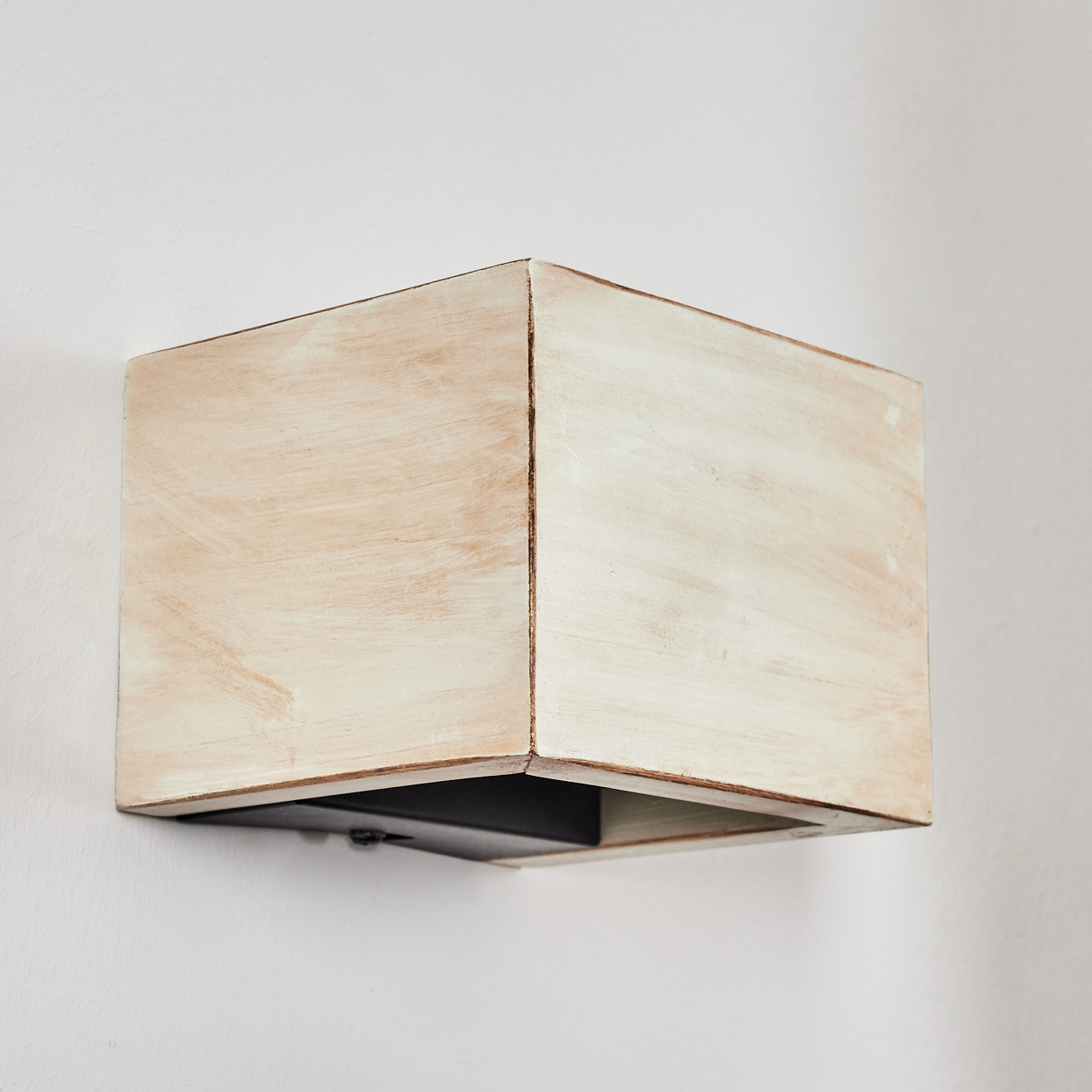 Ein quadratischer, hellfarbiger Holzkasten ist an einer weißen Wand befestigt. Die Unterseite ist schwarz und in Beigetönen bemalt.
