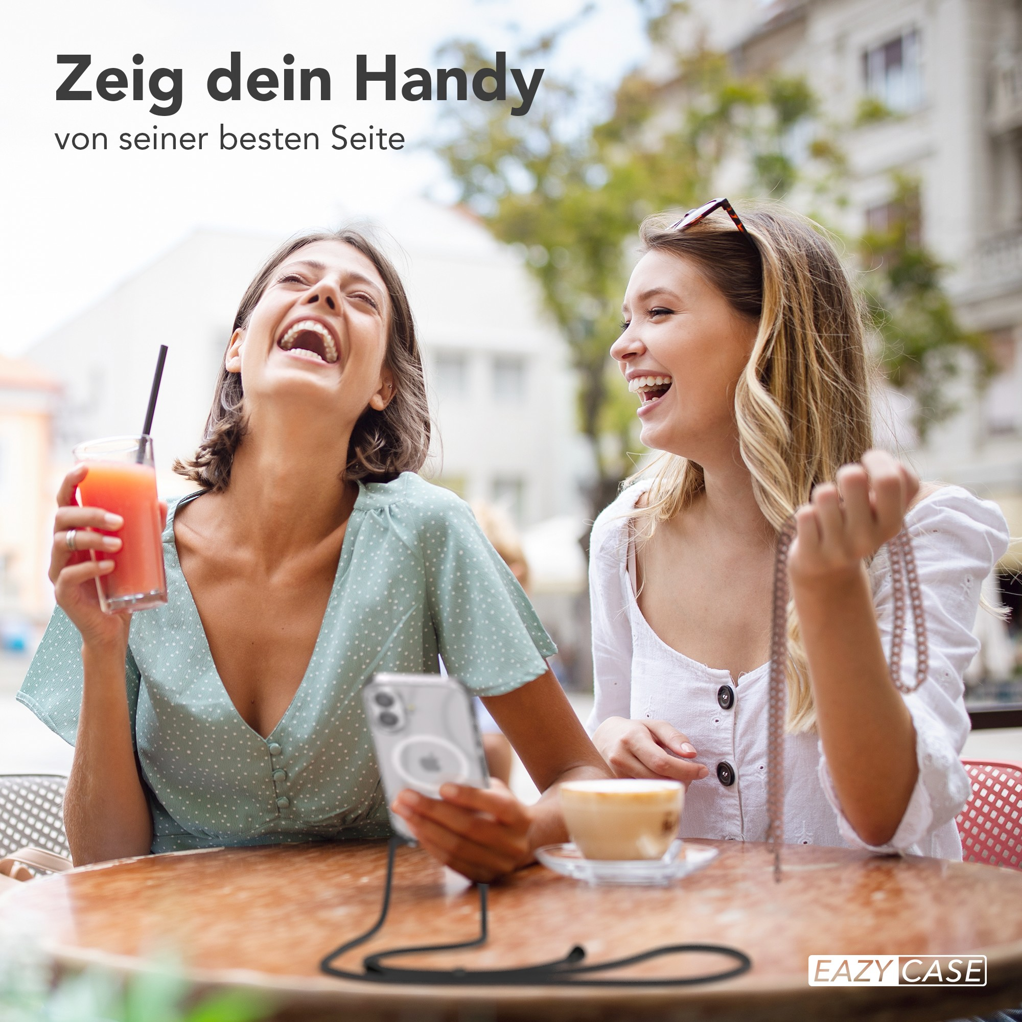 Zwei lachende Frauen an einem Tisch. Eine hält ein Getränk, die andere eine Halskette. Ein Telefon liegt auf dem Tisch.