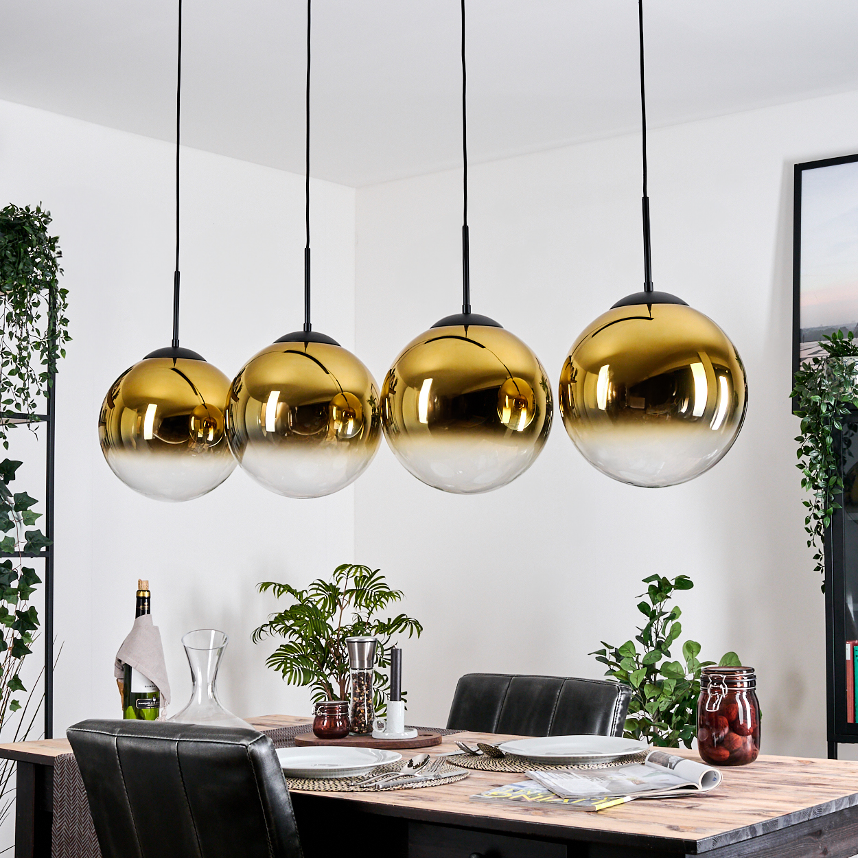 Vier goldene und klare Kugelleuchten hängen über einem Holztisch, der zum Abendessen gedeckt ist. Pflanzen und Dekorationsartikel umgeben den Tisch.