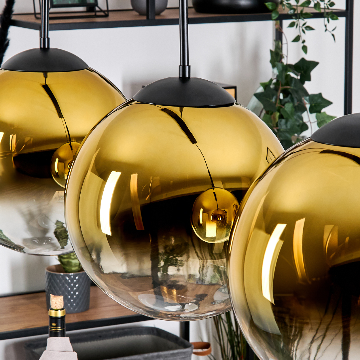 Drei goldene, kugelförmige Pendelleuchten hängen. Im Hintergrund sind eine kleine Pflanze, eine Flasche und eine graue Tonne zu sehen.