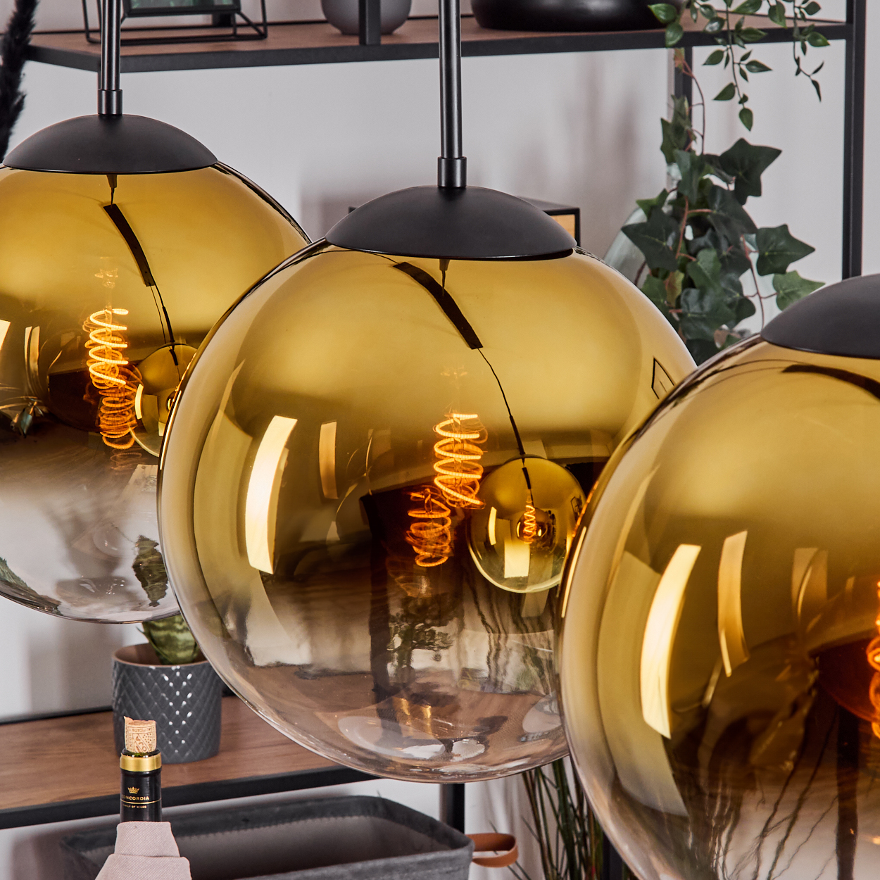 Drei goldene Pendelleuchten mit sichtbaren Glühbirnen. Der Hintergrund umfasst Pflanzen, Regale und eine Weinflasche.