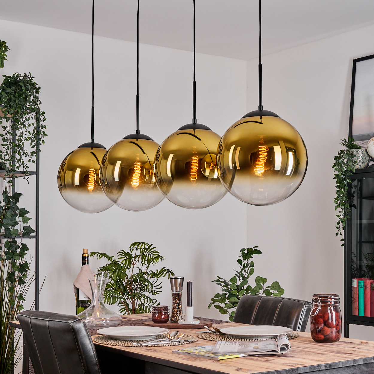 Vier goldene Pendelleuchten über einem Esstisch mit Tellern und Besteck. Pflanzen und ein Glas mit Essen stehen auf dem Tisch.