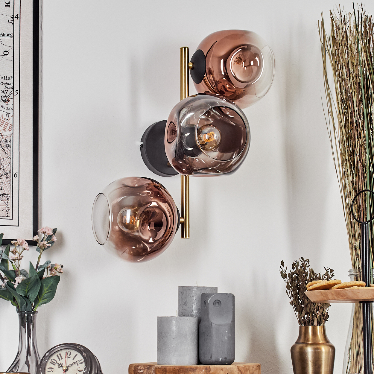 Eine Wandlampe mit drei Glaskugeln. Darunter befinden sich Kerzen und eine Uhr. Eine Vase mit Blumen und eine mit Weizen ist sichtbar.