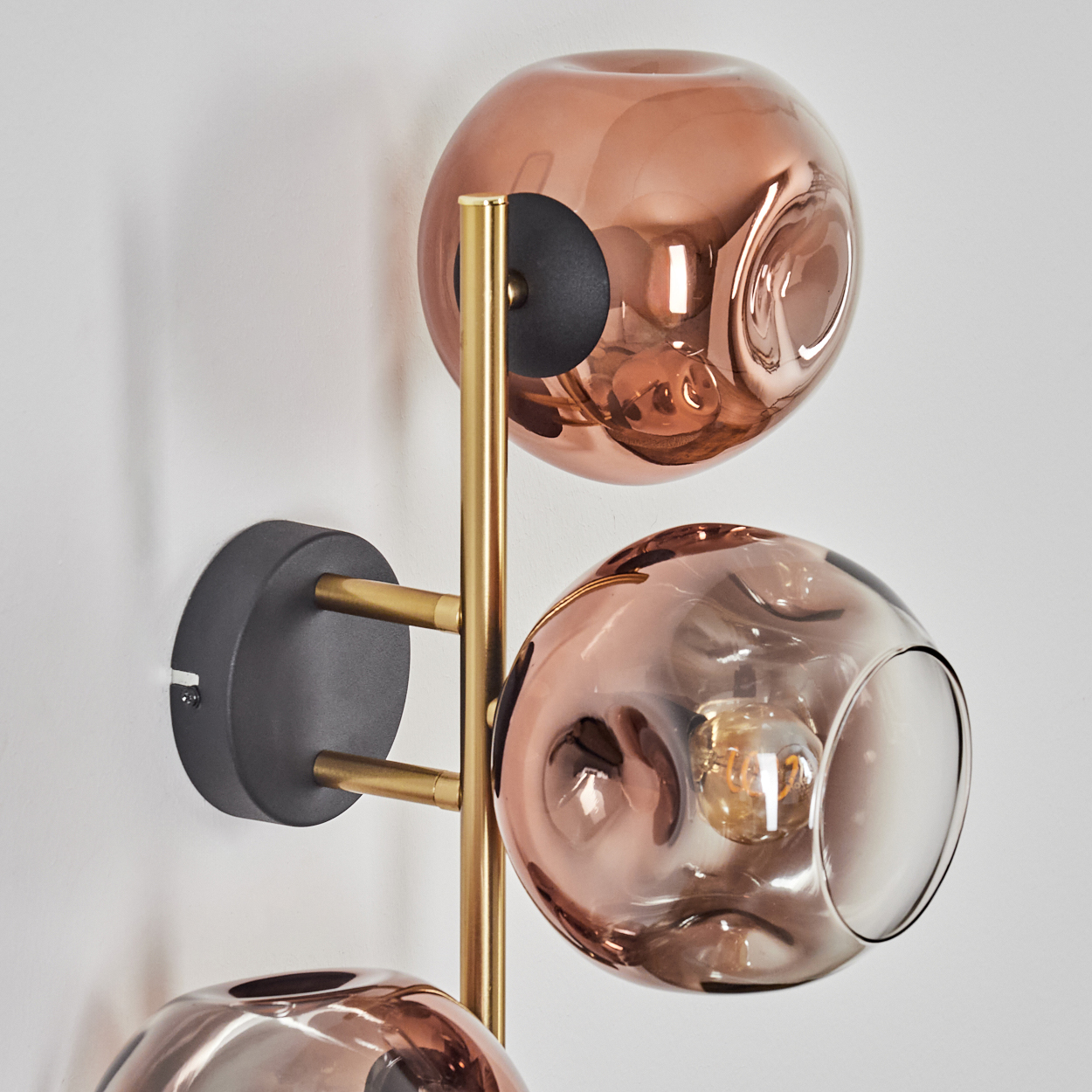 Eine moderne Wandleuchte mit drei kupferfarbenen Glaskugelschirmen, goldenen Stielen und einem schwarzen Sockel an einer weißen Wand.