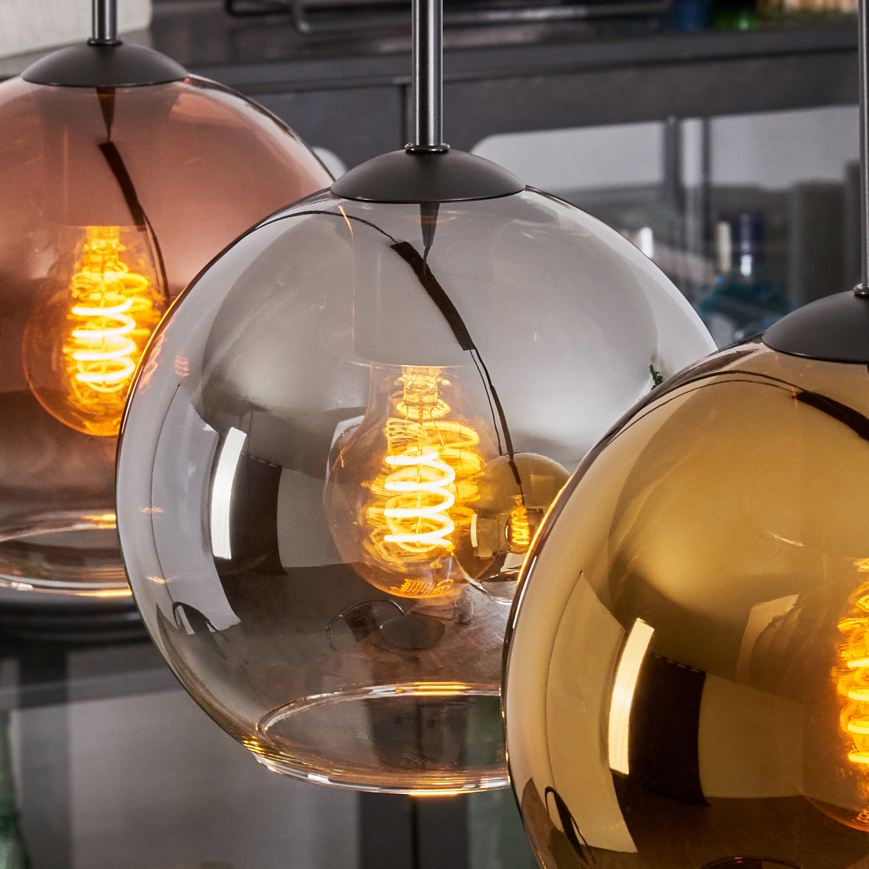 Drei kugelförmige Pendelleuchten. Jede hat einen farbigen Glasschirm und eine glühende Edison-Glühbirne.