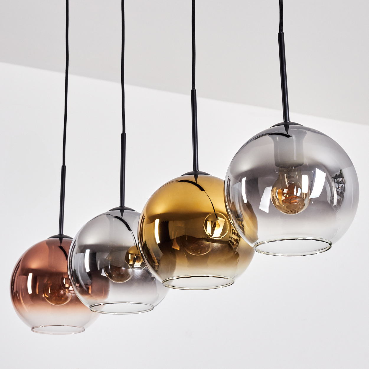 Vier kugelförmige Pendelleuchten. Jede hat eine andere Farbe: Kupfer, Silber, Gold und Grau. Schwarze Kabel hängen von einer weißen Decke.