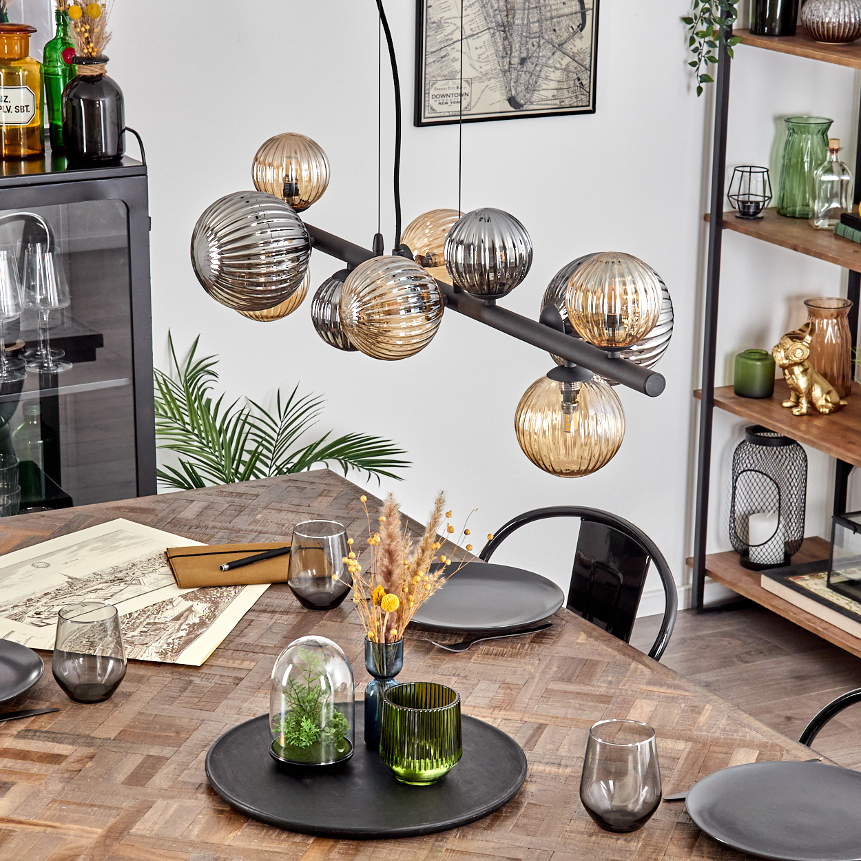 Ein Esstisch mit Tellern, Gläsern und einem Mittelstück. Eine moderne Hängeleuchte hängt darüber. Regale und ein Schrank sind im Hintergrund.