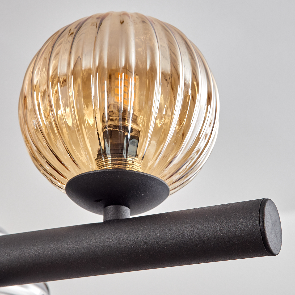 Nahaufnahme einer Leuchte. Sie zeigt eine gerippte, bernsteinfarbene Glaskugel auf einem schwarzen Metallstab vor weißem Hintergrund.