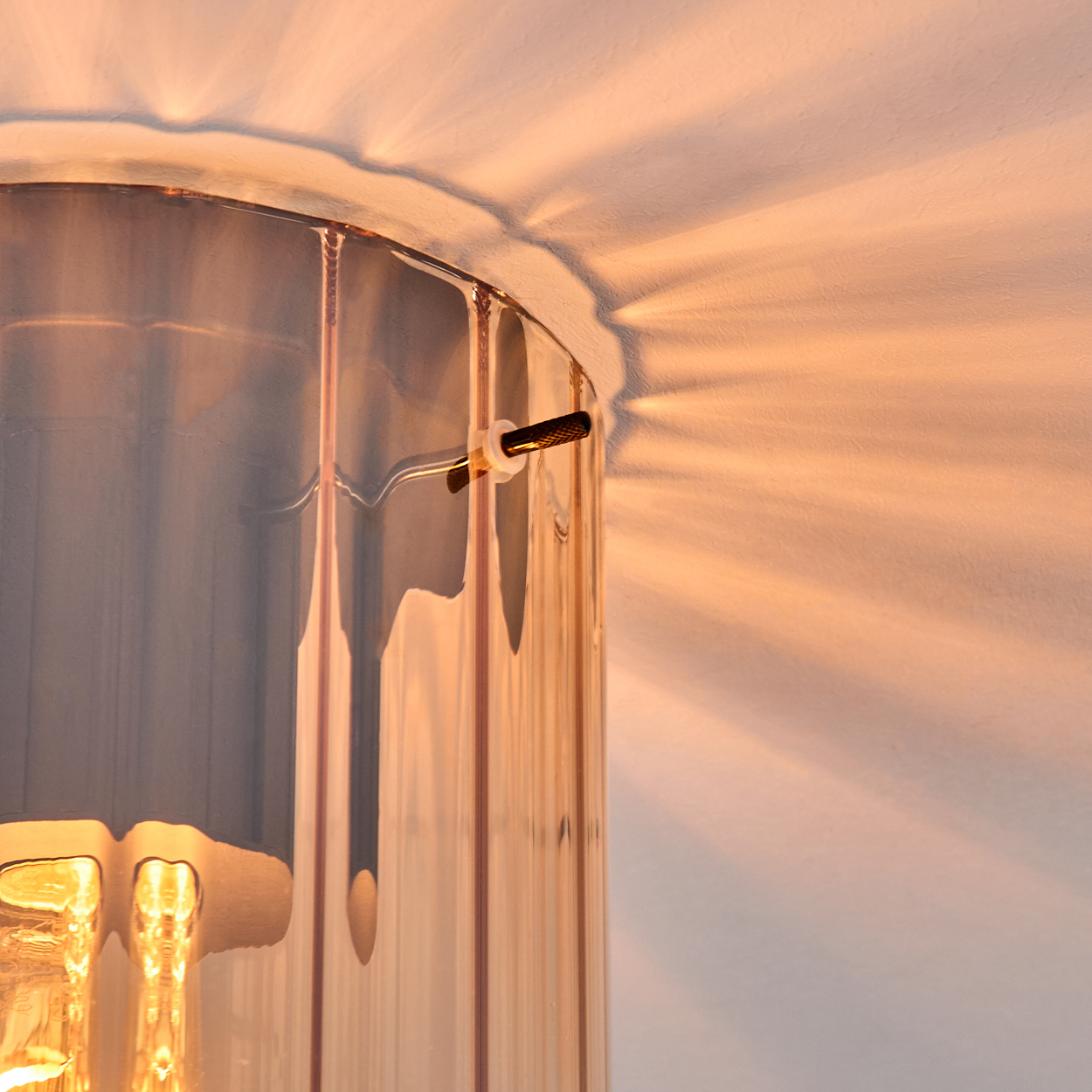 Nahaufnahme einer zylindrischen Glaslampe mit goldenem Licht. Schatten der Lampe werden auf die weiße Wand geworfen.