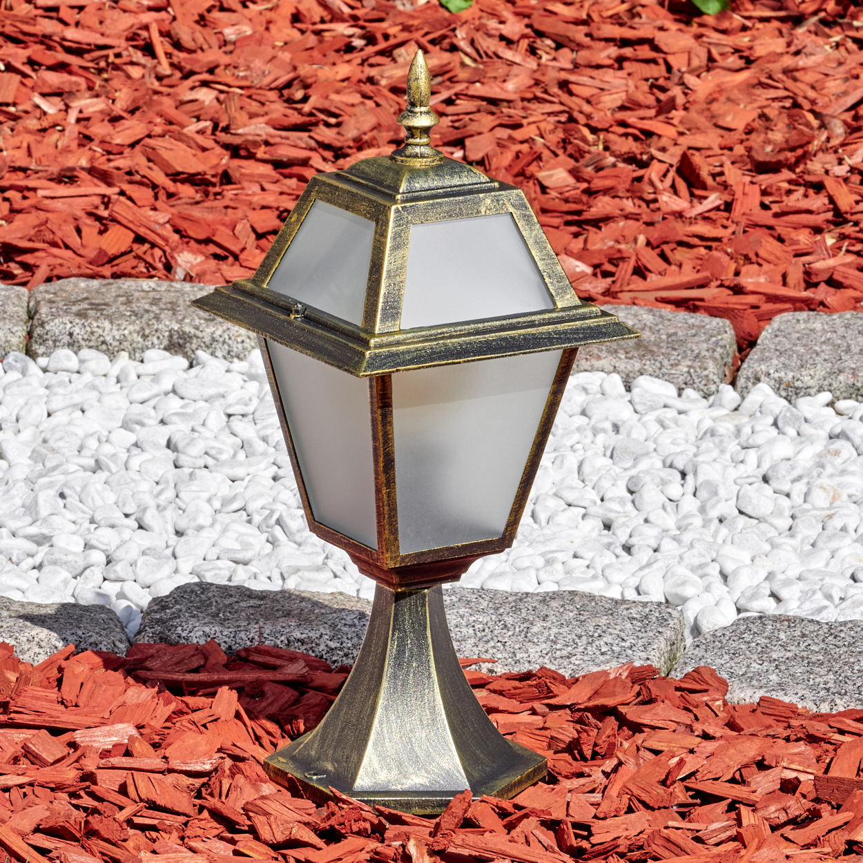 Eine verzierte Außenlampe steht auf roter Mulch. Weiße Steine und graue Steine umranden die Lampe. Die Lampe ist goldfarben und hat Glasseiten.