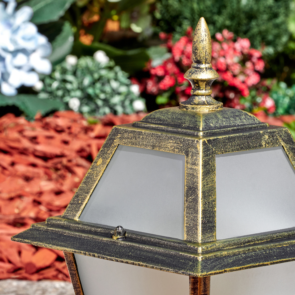 Nahaufnahme einer dekorativen Laterne mit gold-schwarzem Finish. Der Hintergrund hat roten Mulch und bunte Blumen.