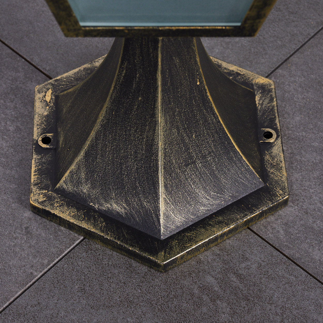 Nahaufnahme eines schwarz-goldenen sechseckigen Lampensockels auf grauen Fliesen. Die Lampe hat ein geometrisches Design und besteht aus Metall.