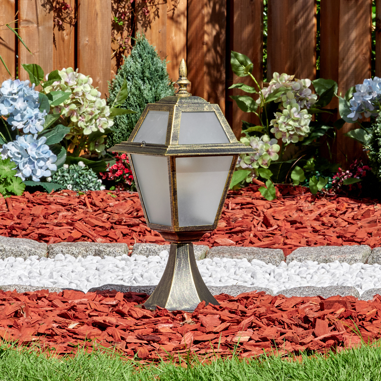 Eine dekorative Außenlampe mit Goldfinish steht in einem Gartenbeet mit roter Mulch, weißen Steinen und Blumen vor einem Holzzaun.