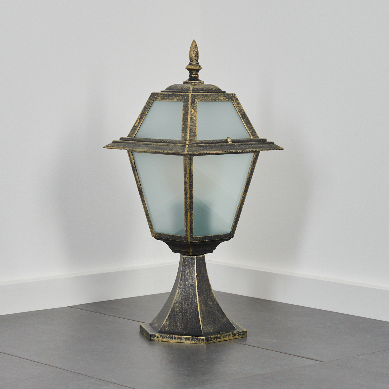 Eine verzierte Außenlampe steht auf einem grauen Fliesenboden. Die Lampe hat ein Milchglasgehäuse und eine Gold-Schwarz-Oberfläche.