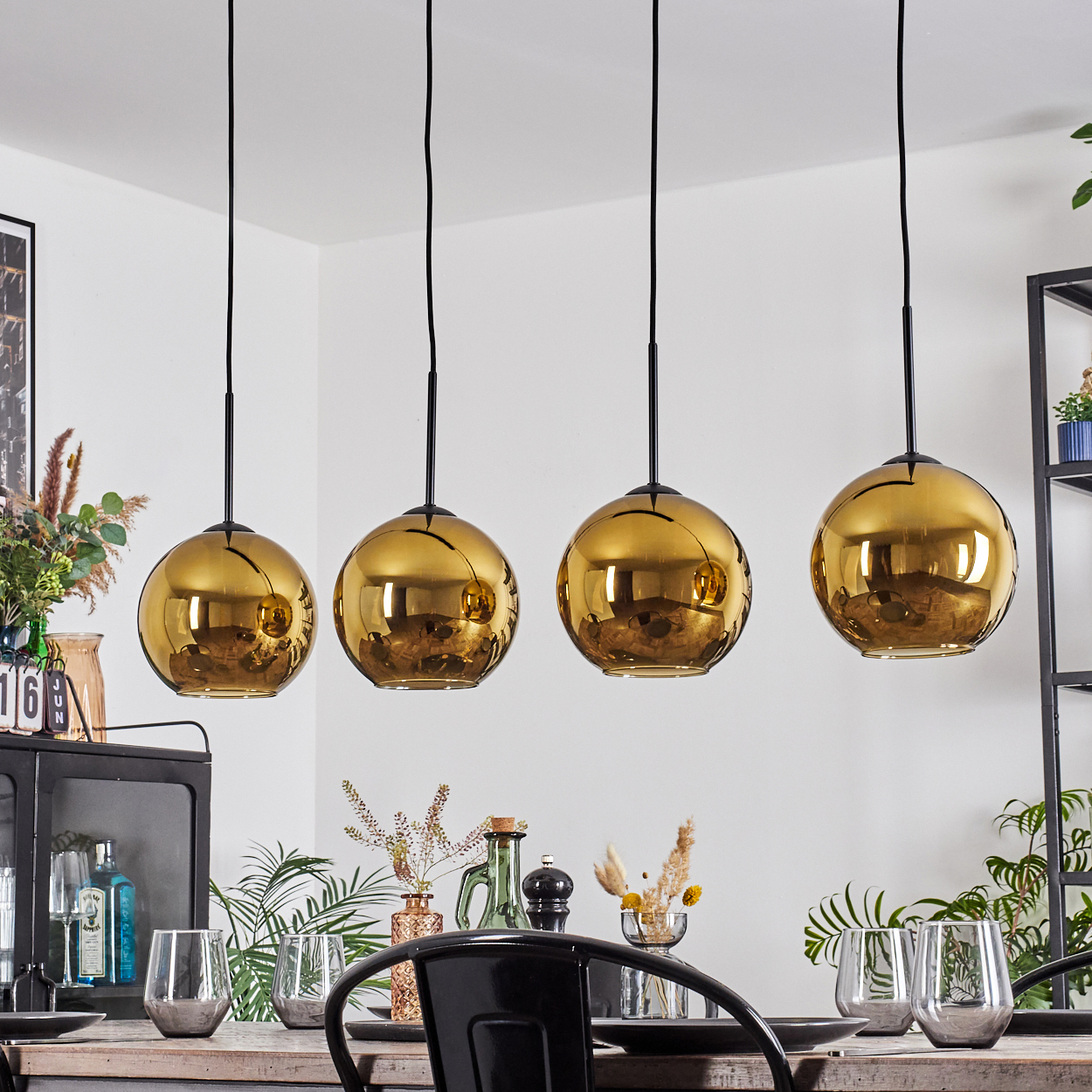 Vier goldene Pendelleuchten über einem Esstisch mit Gläsern, Pflanzen und einem schwarzen Stuhl. Weiße Wand im Hintergrund.