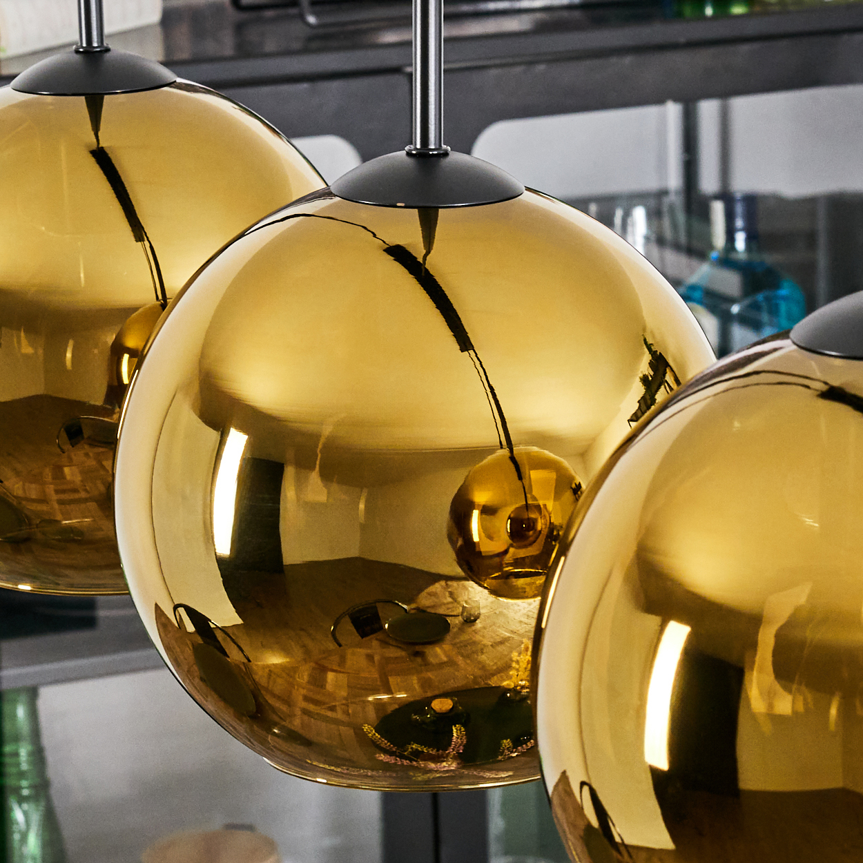 Drei goldene, kugelförmige Pendelleuchten hängen von der Decke. Sie spiegeln die Umgebung wider. Schwarze Metallelemente sind sichtbar.