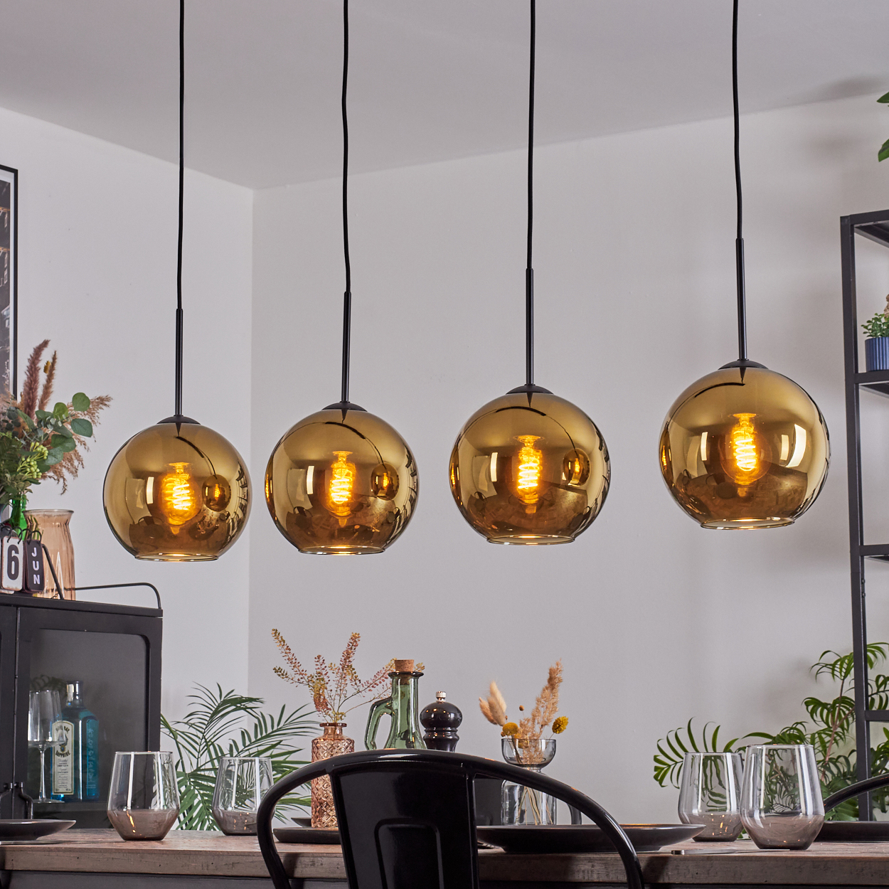 Vier goldene Pendelleuchten hängen über einem Tisch, der mit Gläsern und Dekor gedeckt ist. Hintergrund: Regale und Pflanzen.