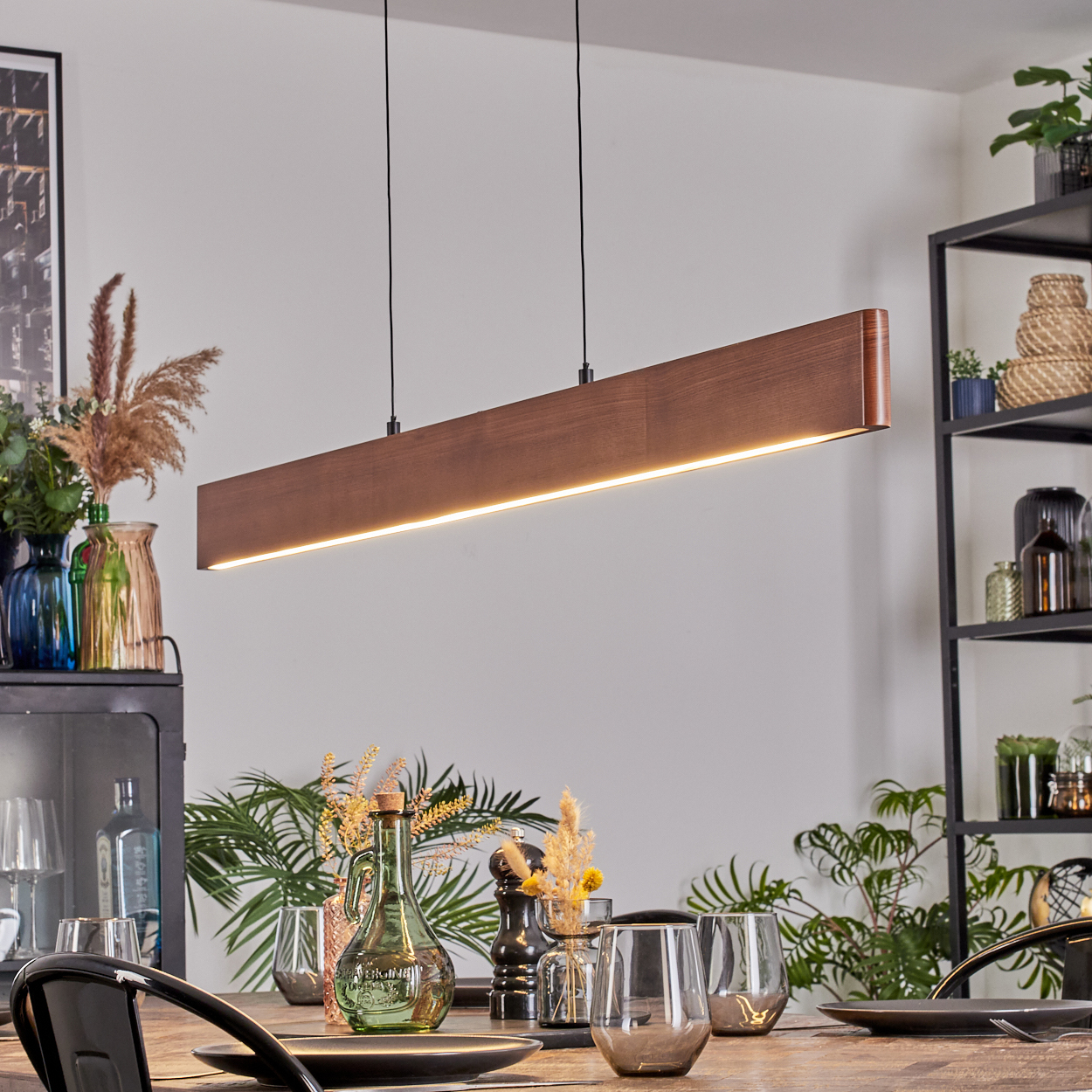 Ein modernes Esszimmer mit einer rechteckigen Holzleuchte. Tisch mit Gläsern und Tellern gedeckt. Vasen und Pflanzen sind vorhanden.