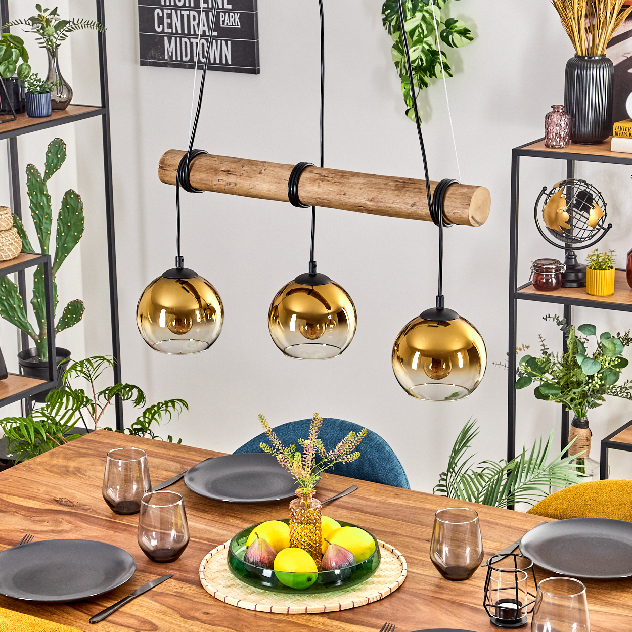 Esstisch mit goldenen Pendelleuchten. Teller, Gläser und eine Obstschale stehen auf dem Holztisch. Pflanzen und Regale im Hintergrund.