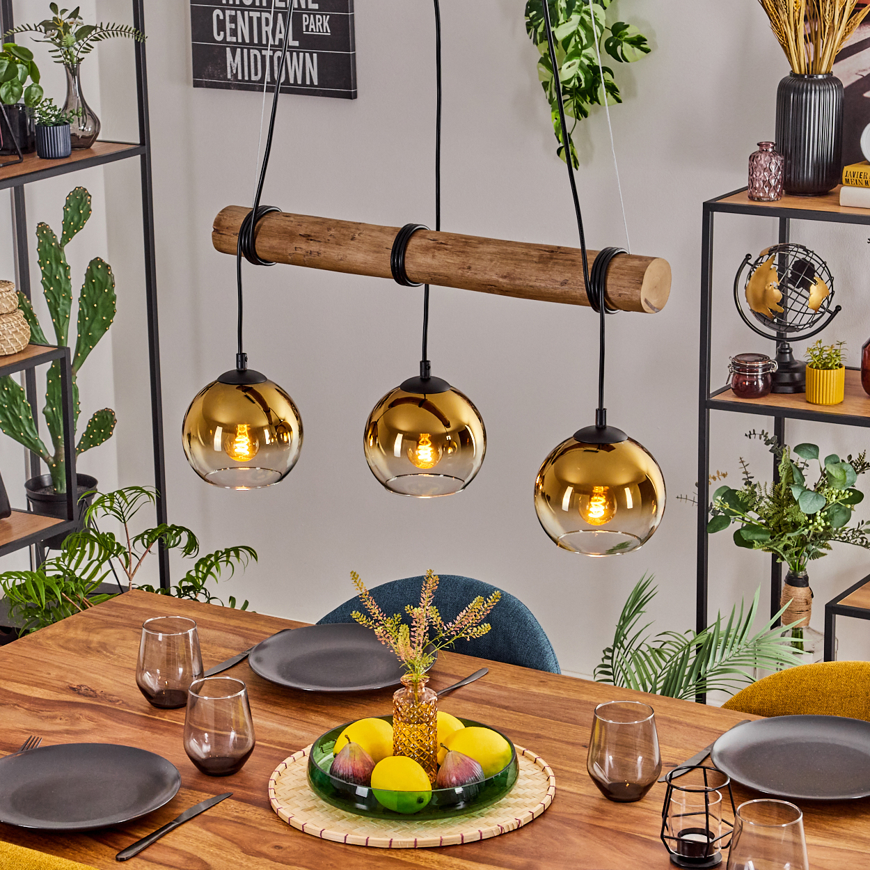 Esstisch mit goldenen Pendelleuchten. Früchte und Gläser stehen auf dem Tisch. Pflanzen und Regale im Hintergrund.
