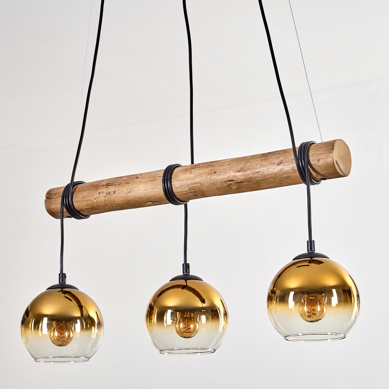 Eine moderne Pendelleuchte mit drei goldenen Glaskugelschirmen, die an einem Holzbalken hängen. Schwarze Drähte und ein weißer Hintergrund.