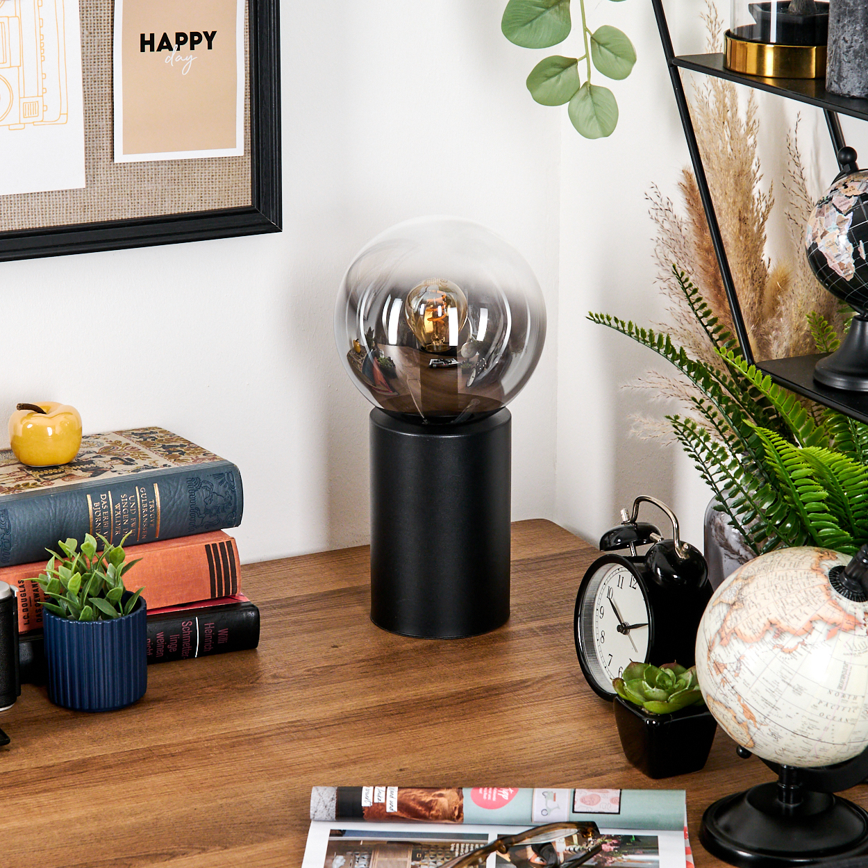 Ein Schreibtisch mit Lampe, Büchern, Uhr und Globus. Im Hintergrund Regale und Pflanzen. Ein Bilderrahmen mit "Happy day"-Schild ist an der Wand.