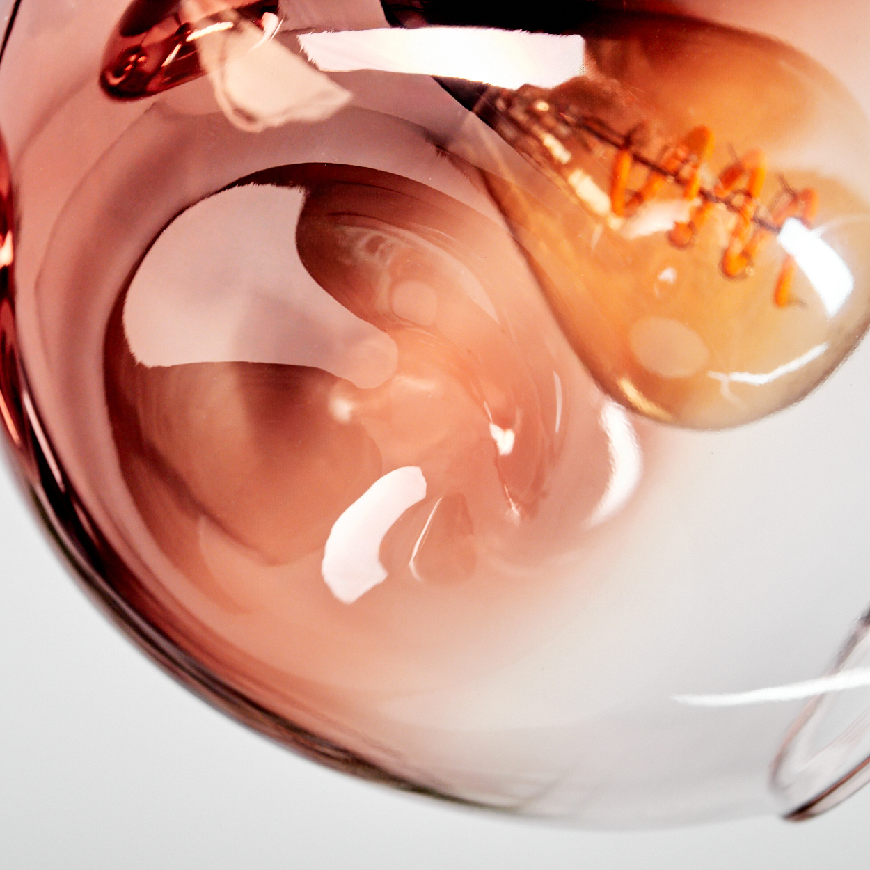 Nahaufnahme einer Glühbirne mit sichtbarem Glühfaden. Die Glühbirne hat einen rosa-orangen Farbton und reflektiert Licht. Weißer Hintergrund.