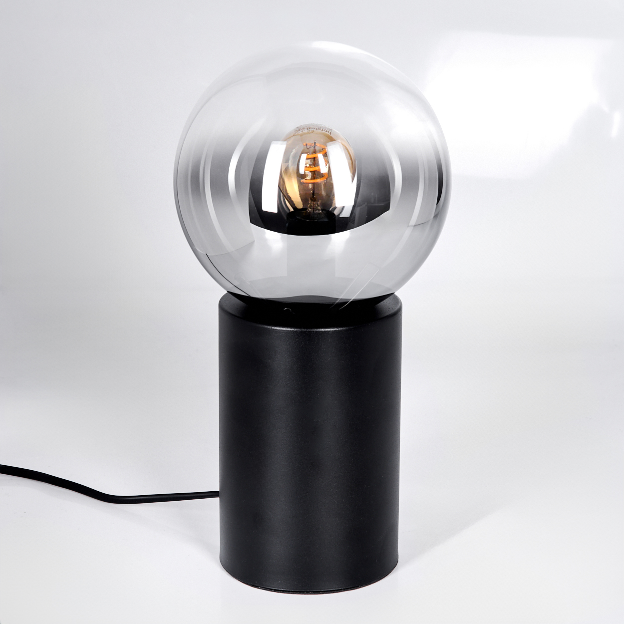 Eine moderne Lampe mit schwarzem zylindrischen Sockel und einem klaren, kugelförmigen Schirm. Im Schirm befindet sich eine leuchtende Glühbirne.