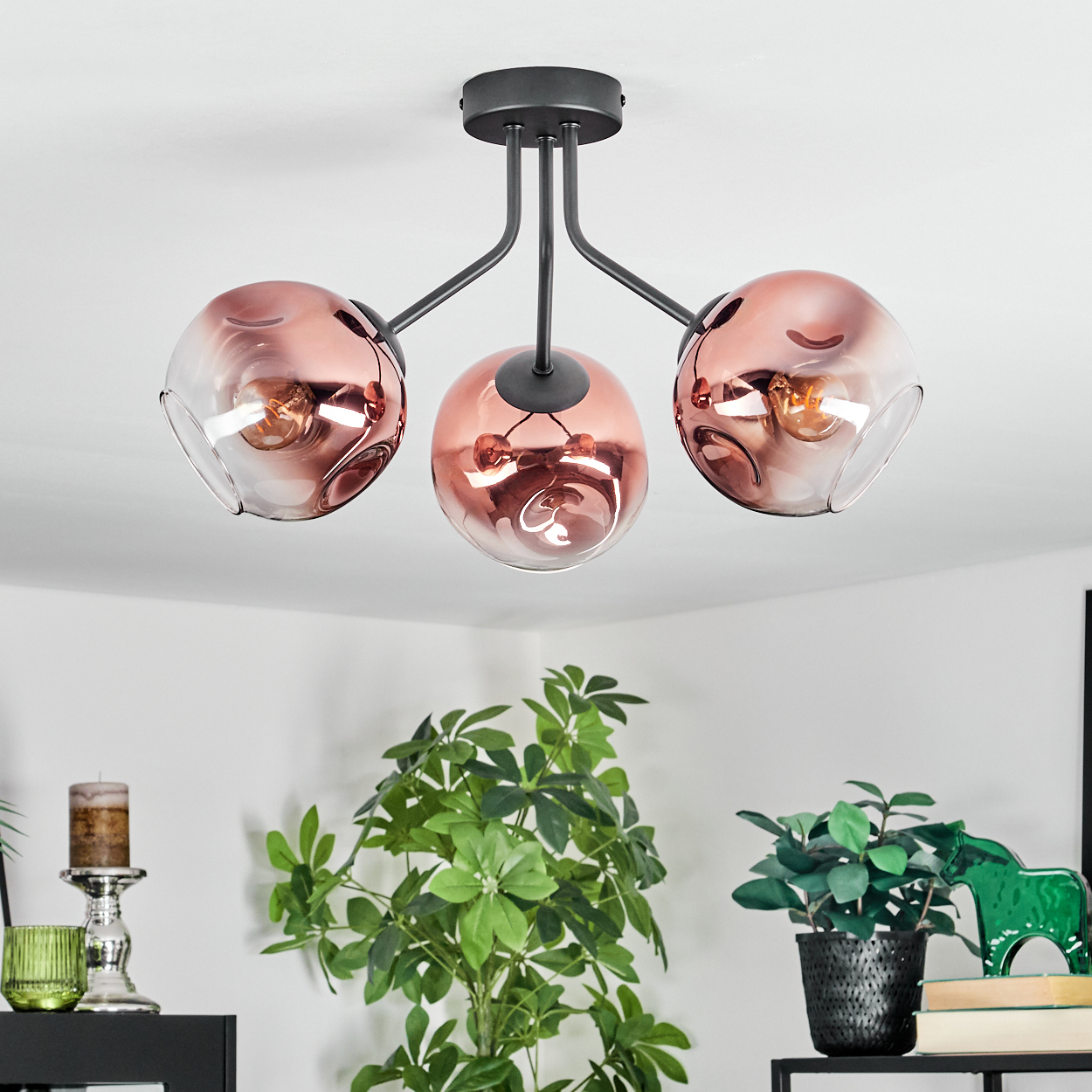 Eine Deckenleuchte mit drei roségoldenen Glaskugeln. Darunter grüne Pflanzen und eine Kerze. Weißer Wandhintergrund.