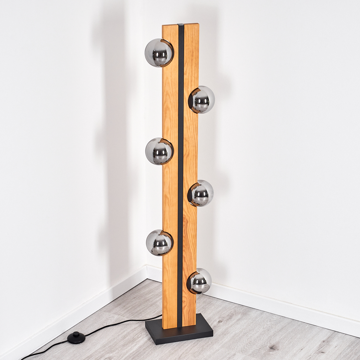 Eine moderne Stehlampe mit Holzrahmen und sechs kugelförmigen Chromleuchten. Sie steht auf einem schwarzen Sockel. Der Hintergrund ist eine weiße Wand und ein Holzboden.