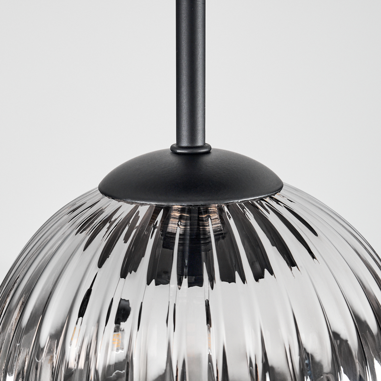 Nahaufnahme einer Glas-Pendelleuchte. Sie hat einen gerippten, halbkugelförmigen Schirm und eine schwarze Metallhalterung. Der Hintergrund ist weiß.
