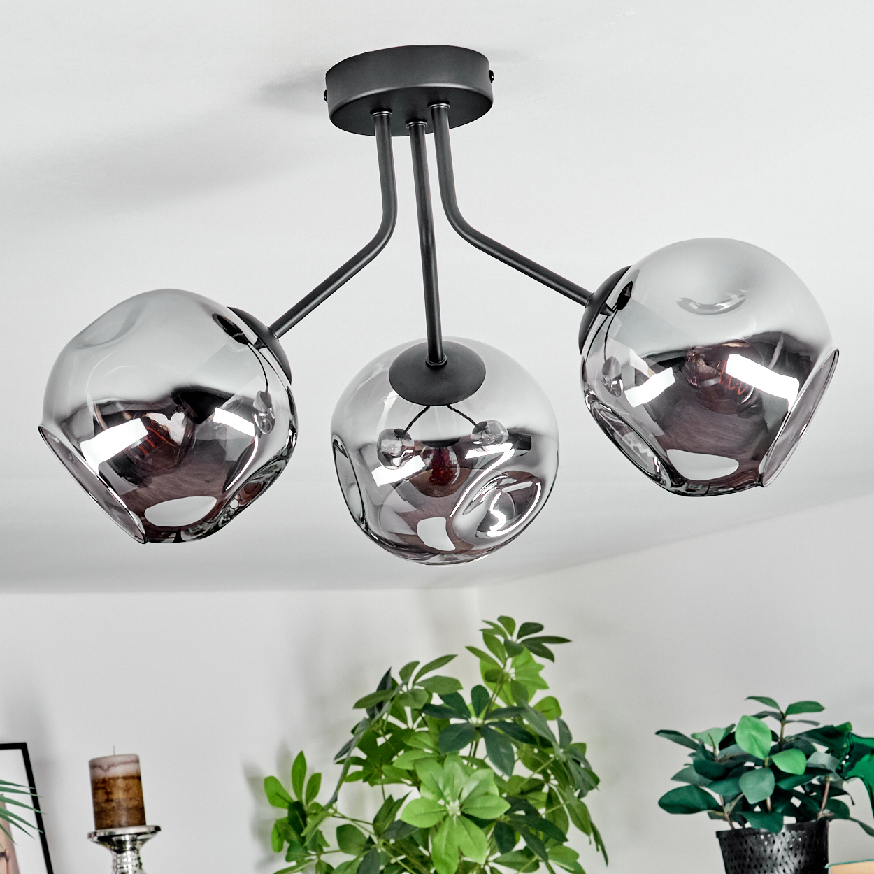 Eine moderne Deckenlampe mit drei rauchigen Glasschirmen. Im Vordergrund grüne Pflanzen.