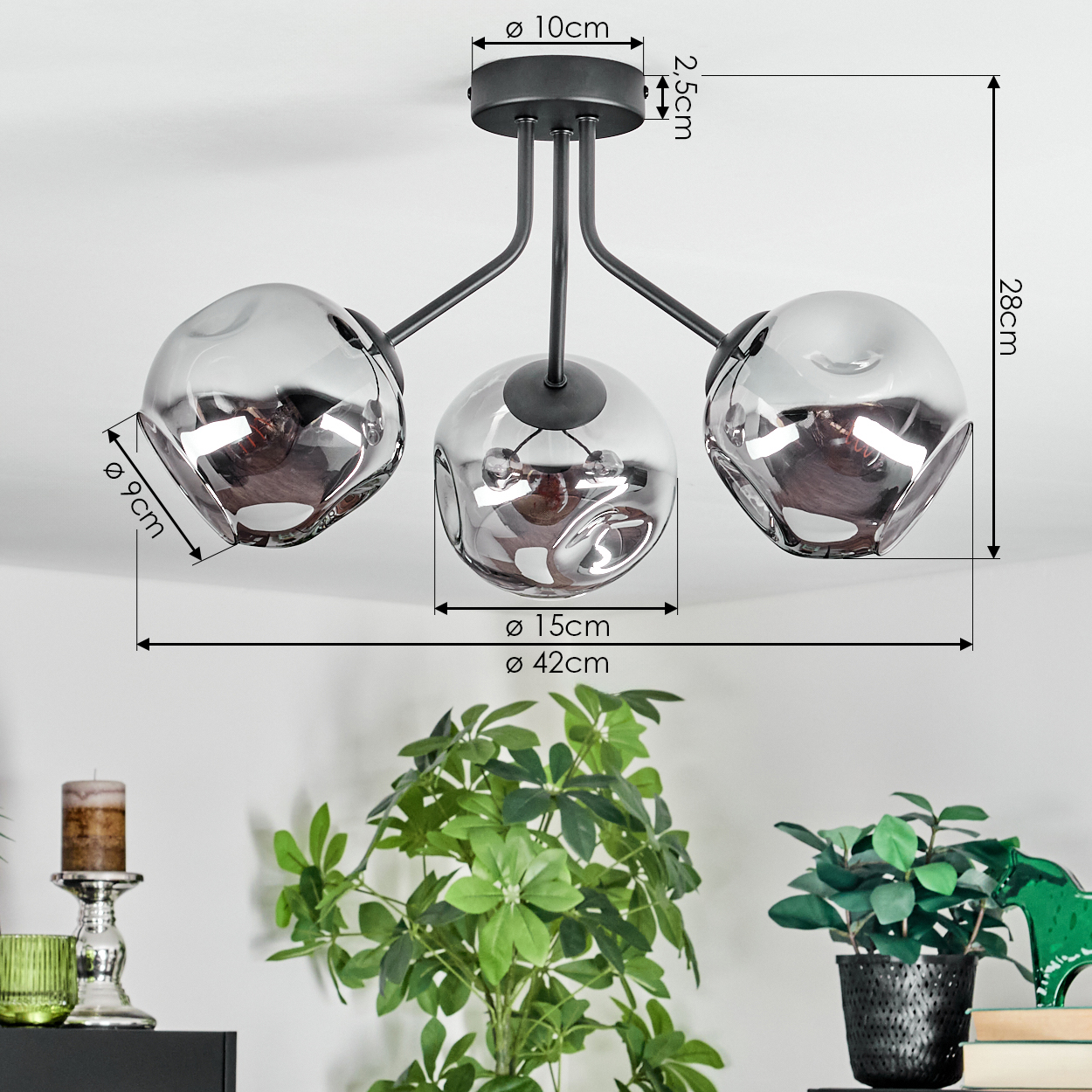 Eine moderne Deckenlampe mit drei Glasschirmen. Grüne Pflanzen und ein Kerzenhalter sind im Vordergrund, mit Maßangaben.
