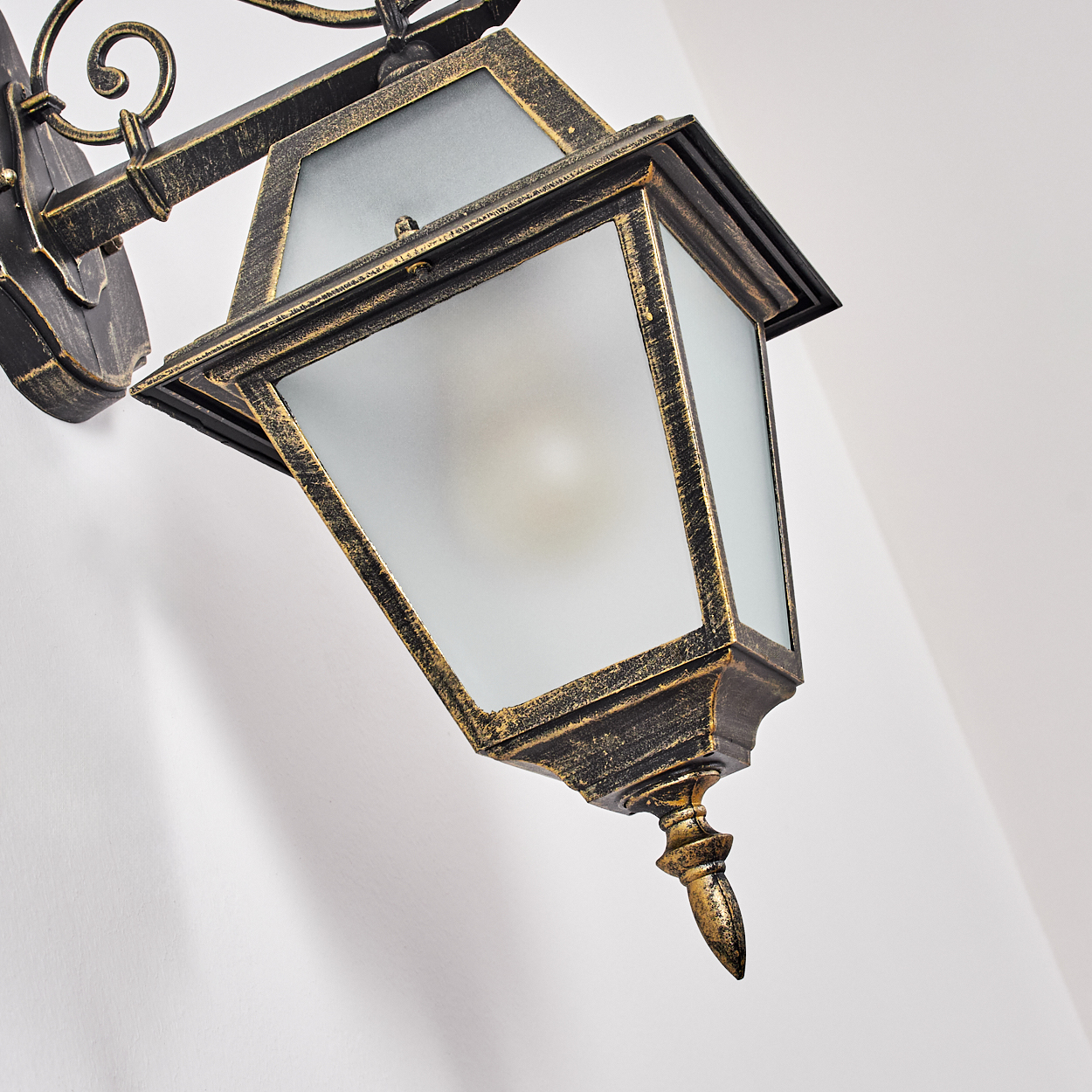 Eine verzierte, bronzefarbene Wandlampe mit einem Schirm aus Milchglas. Sie ist an einer weißen Wand montiert, abgewinkelt, mit einer dekorativen Halterung.