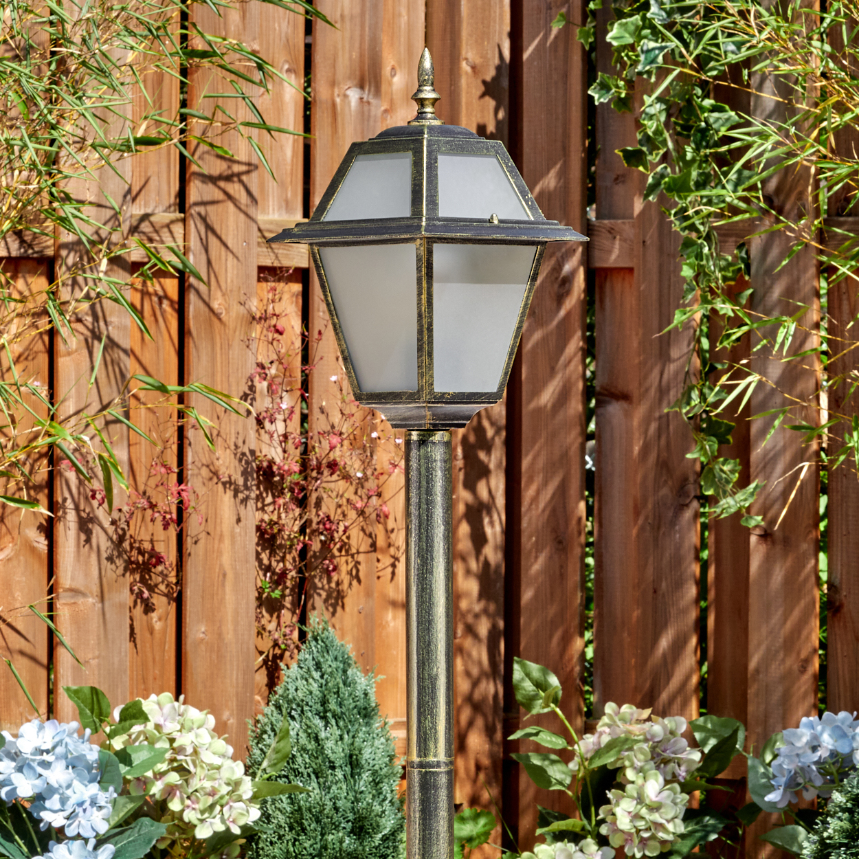 Eine dekorative Außenlampe steht vor einem Holzzaun, umgeben von Pflanzen und Blumen in einem Garten.