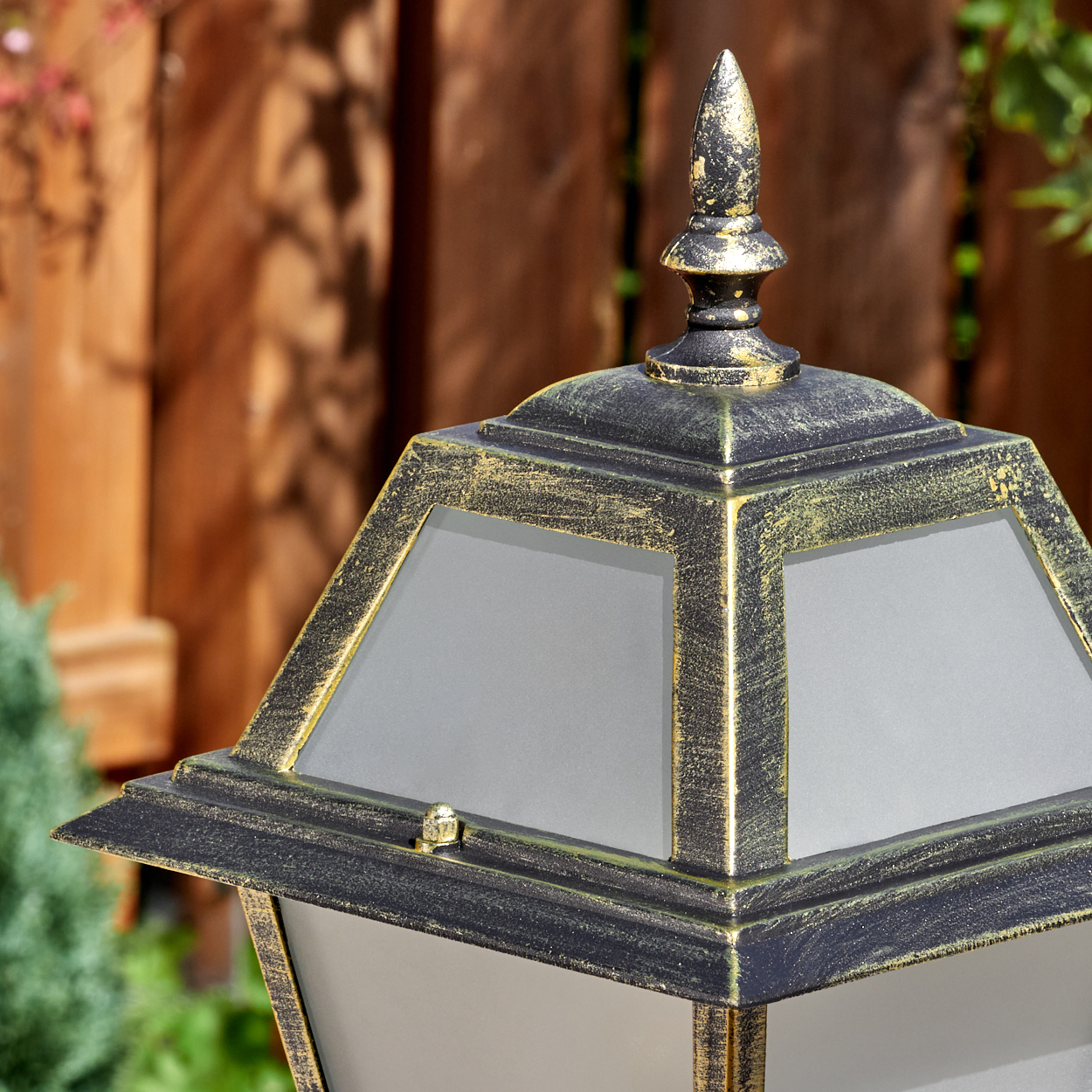 Nahaufnahme einer schwarz-goldenen Außenlampe. Ein Holzzaun und grünes Laub sind im unscharfen Hintergrund.