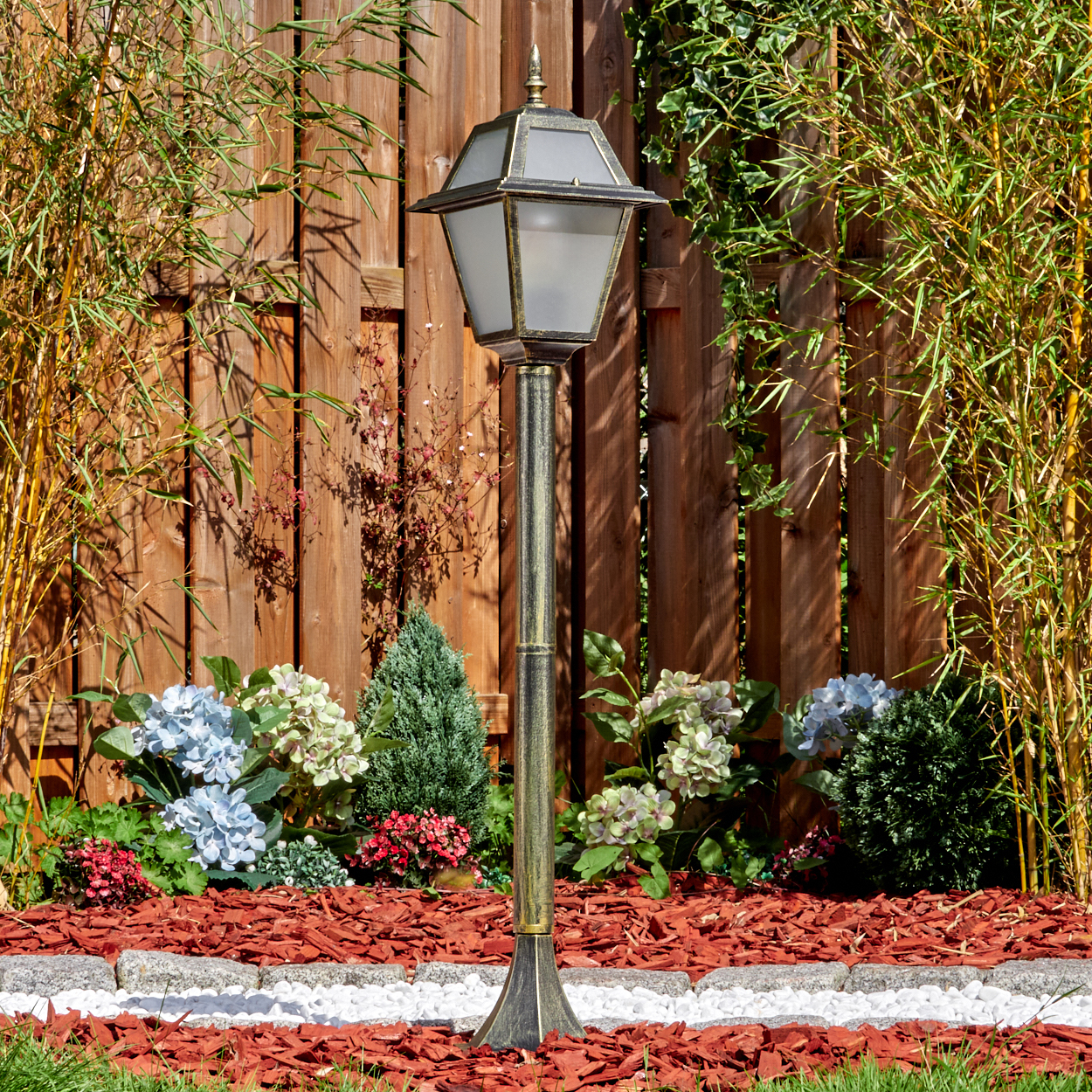 Eine dekorative Gartenlampe steht vor einem Holzzaun, umgeben von Pflanzen und rotem Mulch. Bambus befindet sich auf beiden Seiten.
