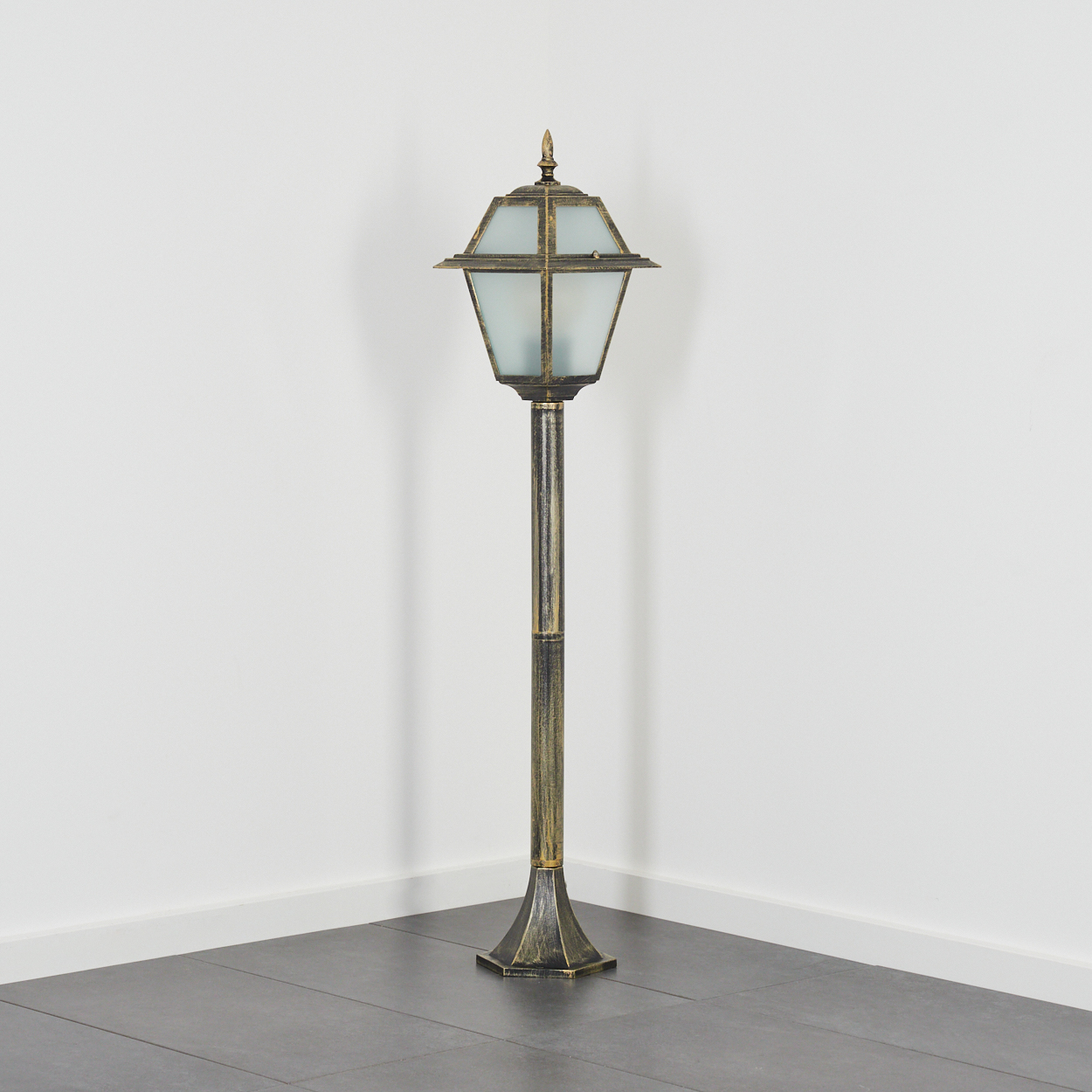 Eine antike Außenlampe. Sie hat einen goldfarbenen Metallrahmen mit einem Schirm aus Milchglas. Sie steht auf einem dunklen Fliesenboden.