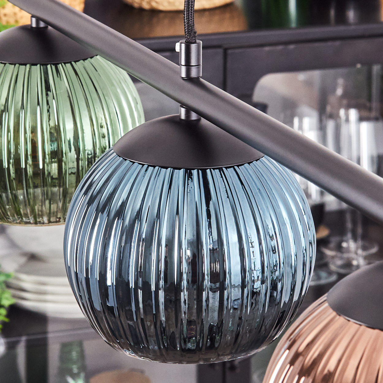 Nahaufnahme eines modernen Kronleuchters mit drei kugelförmigen Glasschirmen in Grün, Blau und Kupfer. Sie hängen an einem dunklen Metallrahmen.