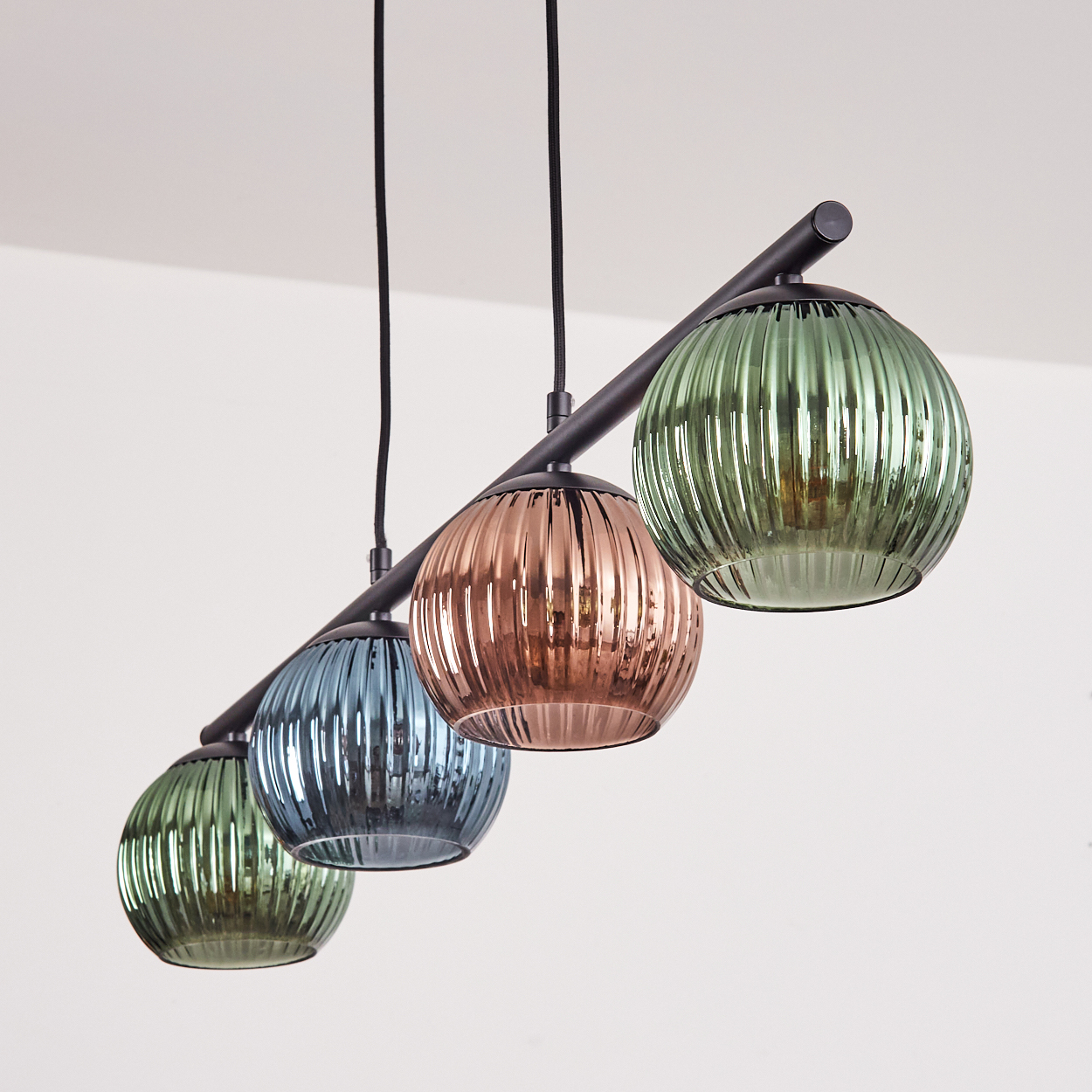 Vier bunte Glas-Pendelleuchten hängen an einer schwarzen Stange. Die Kugeln sind grün, blau, kupferfarben und grün, vor weißem Hintergrund.