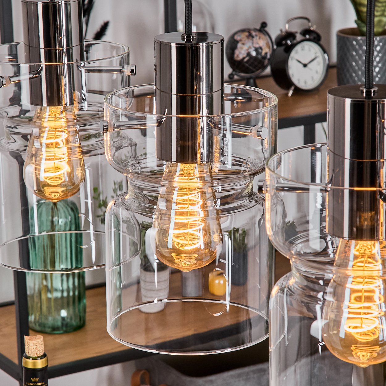 Nahaufnahme von drei Glas-Pendelleuchten mit Edison-Glühbirnen. Im Hintergrund ein Regal mit Deko: Uhr, Globus und Pflanzen.